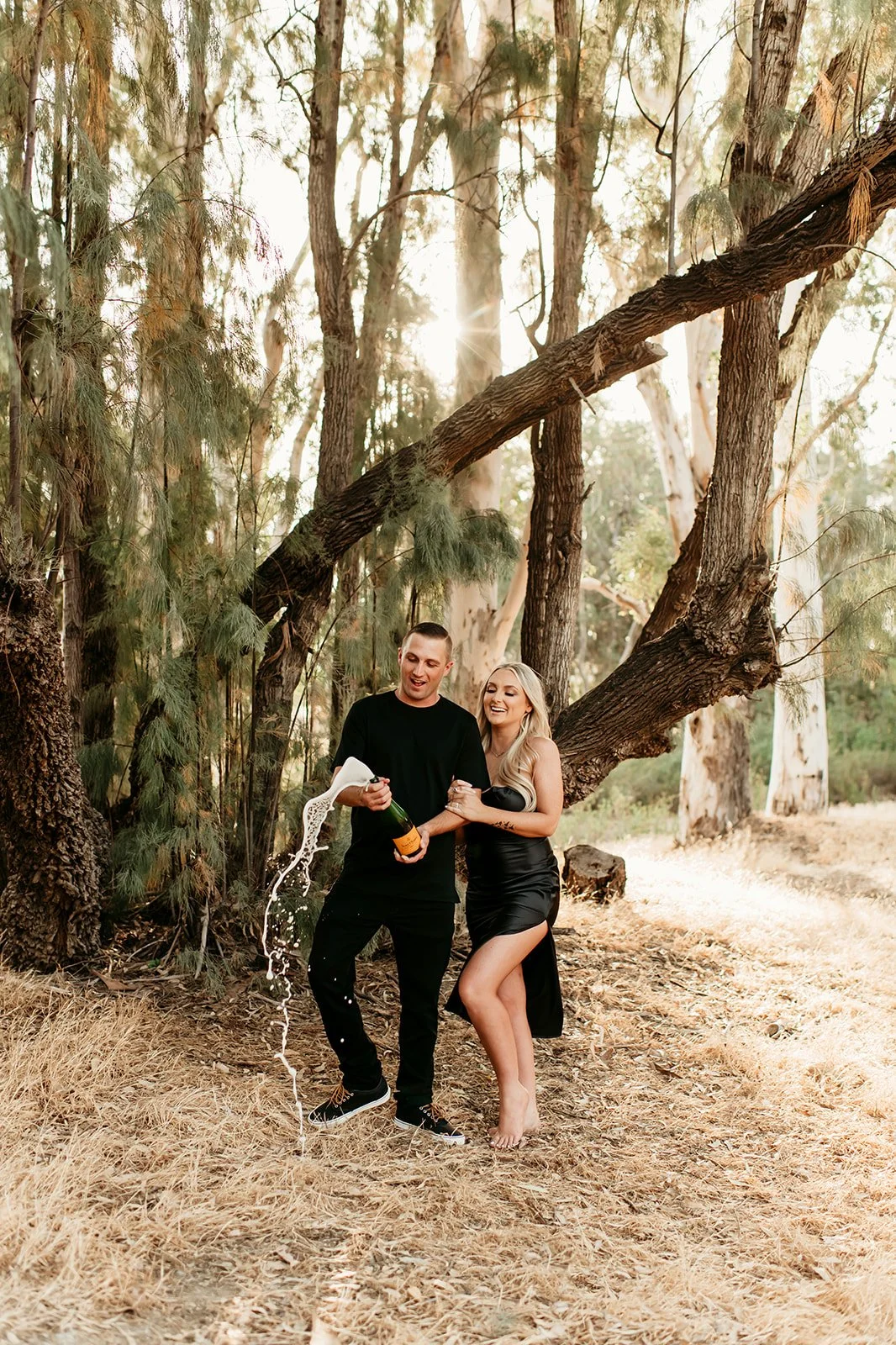 A couple celebrating in a wooded area, opening a bottle of champagne with some spilling out, sunlight filtering through the trees.