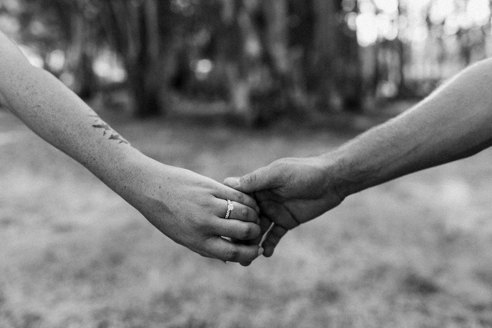 Close-up of two people holding hands, with a focus on one person's hand wearing an engagement ring. The background is blurred, showing a natural outdoor setting in black and white.