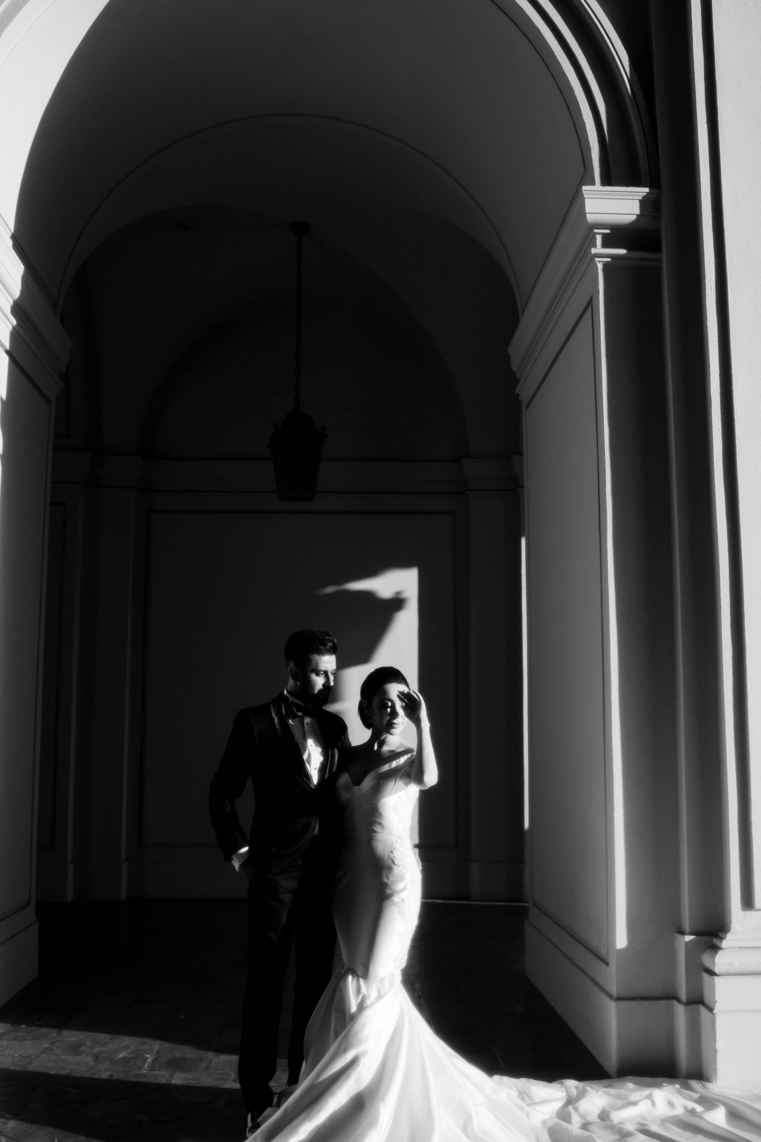 A black and white photo of a man and woman dressed in formal wedding attire, standing in an arched doorway with sunlight casting shadows.
