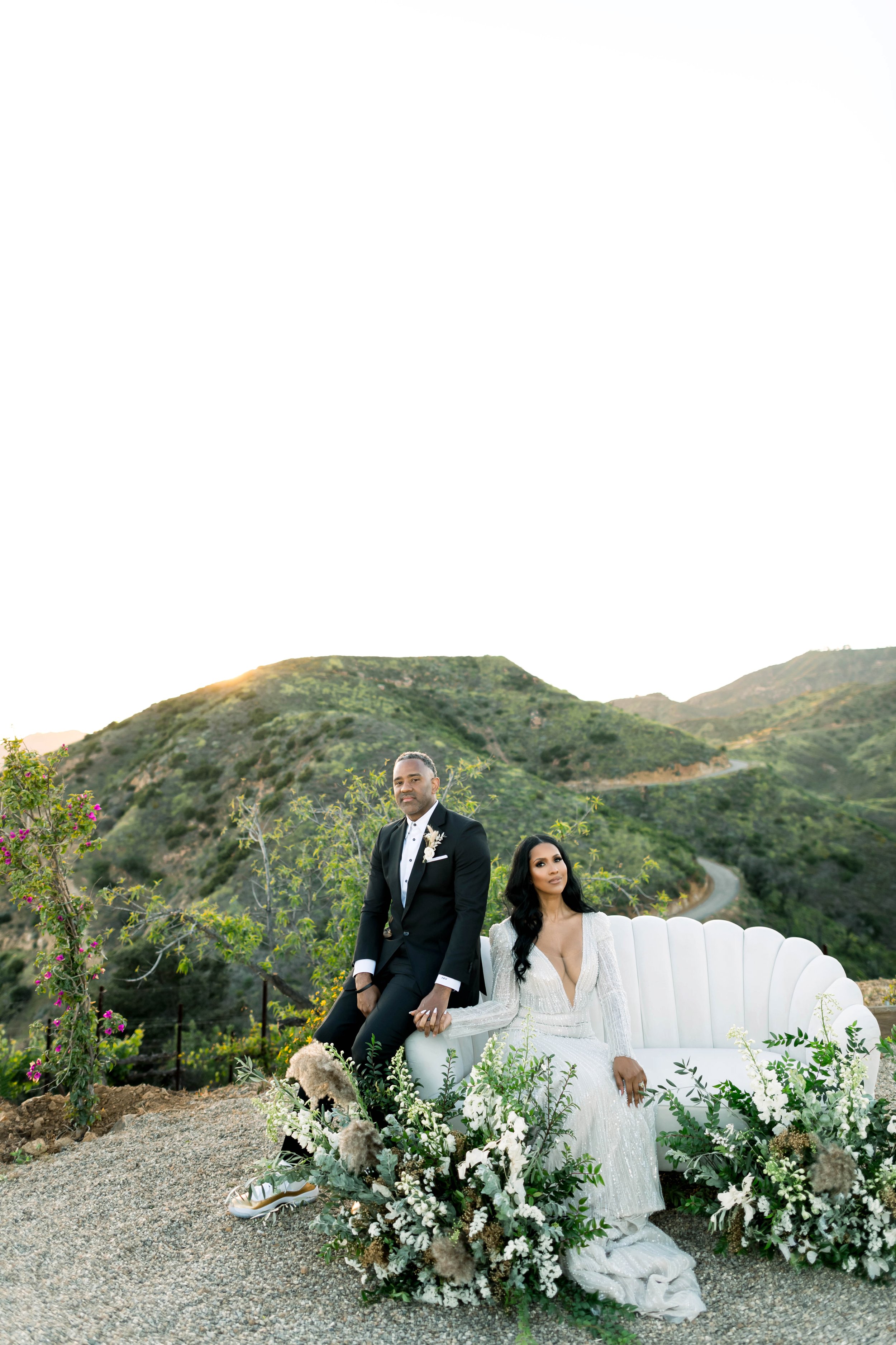 A couple in wedding attire sitting on a white sofa outdoors with mountain scenery in the background. The man wears a black tuxedo with a white shirt and boutonniere, and the woman wears a white gown with long dark hair. Surrounding them are white and