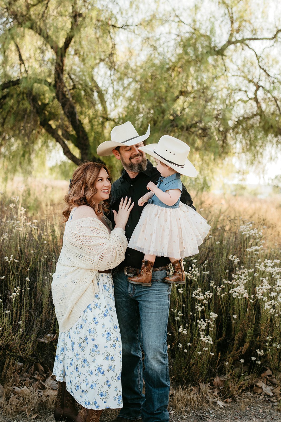 A family of three, a woman, a man, and a young girl, enjoying time outdoors in a grassy, flowery field under a large tree. The woman has long, wavy brown hair and is wearing a white dress with blue floral patterns and a cream-colored open-knit sweate