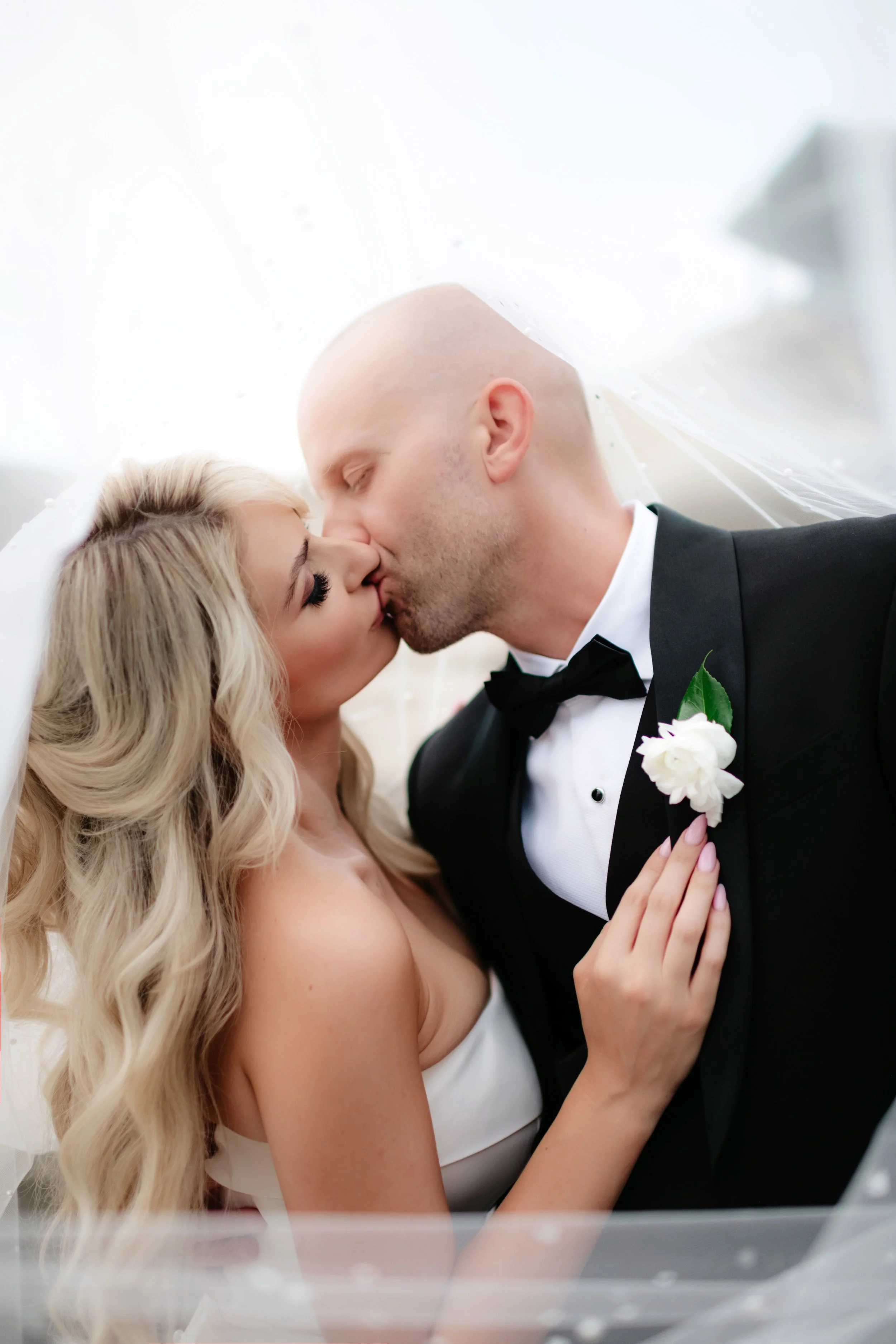 A bride and groom sharing a kiss under a veil on their wedding day, with the bride having long blonde hair and the groom in a tuxedo.