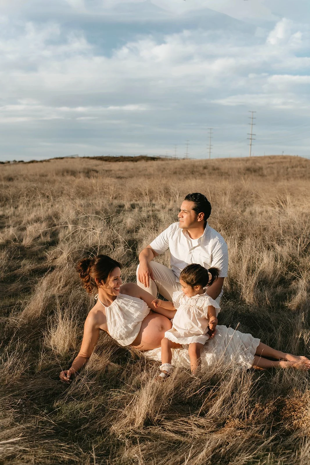 A family of three sitting on a grassy field during sunset. The pregnant woman is lying down in a white dress, the man is sitting beside her in a white shirt, and a young girl in a light dress is sitting on the woman’s lap.