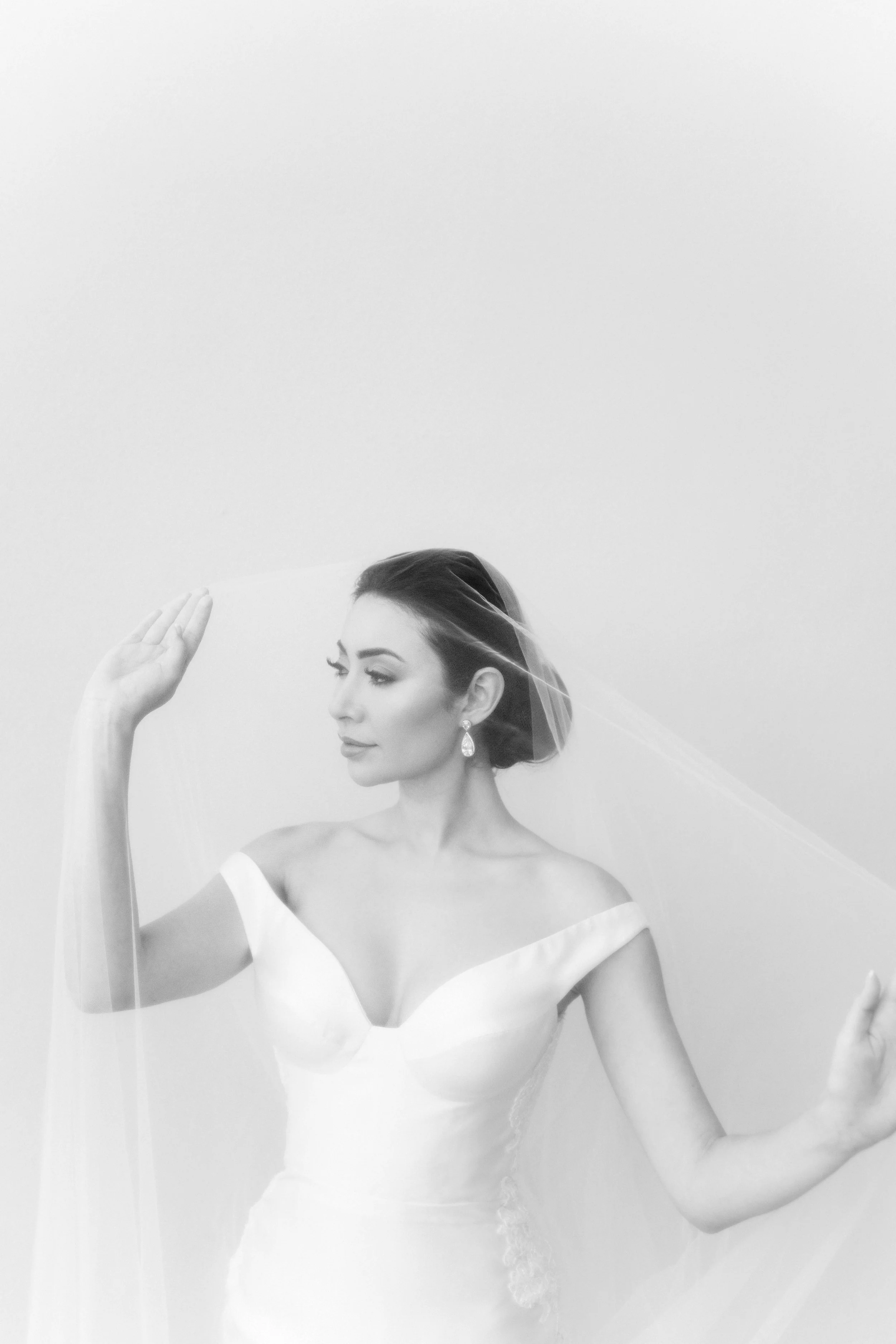 Black and white portrait of a bride in a wedding dress, with her eyes closed and her hand raised to her hair, wearing earrings and a veil.