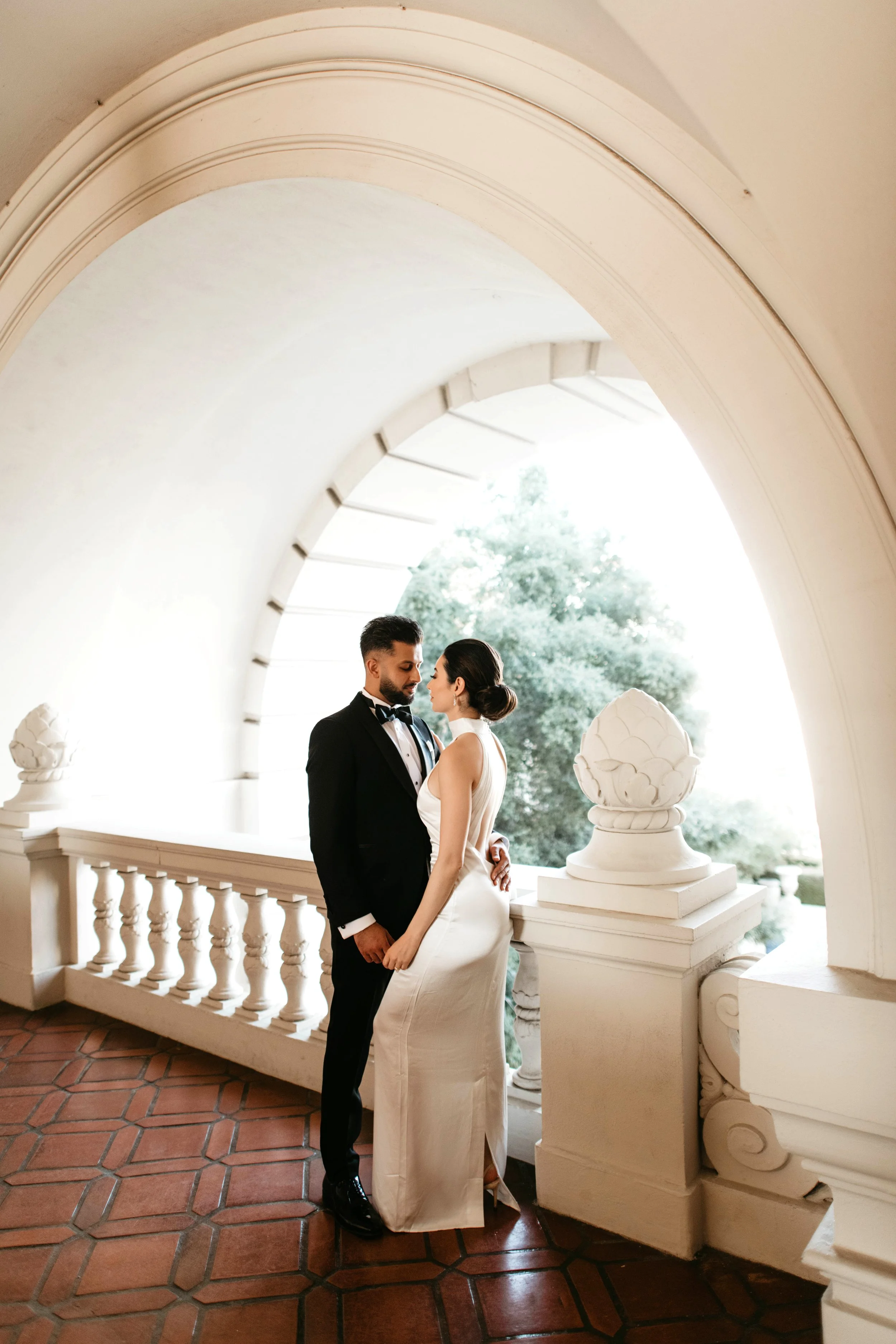 A bride and groom standing close together on a balcony with white balustrades and urn decorations, under a large archway with trees visible in the background.