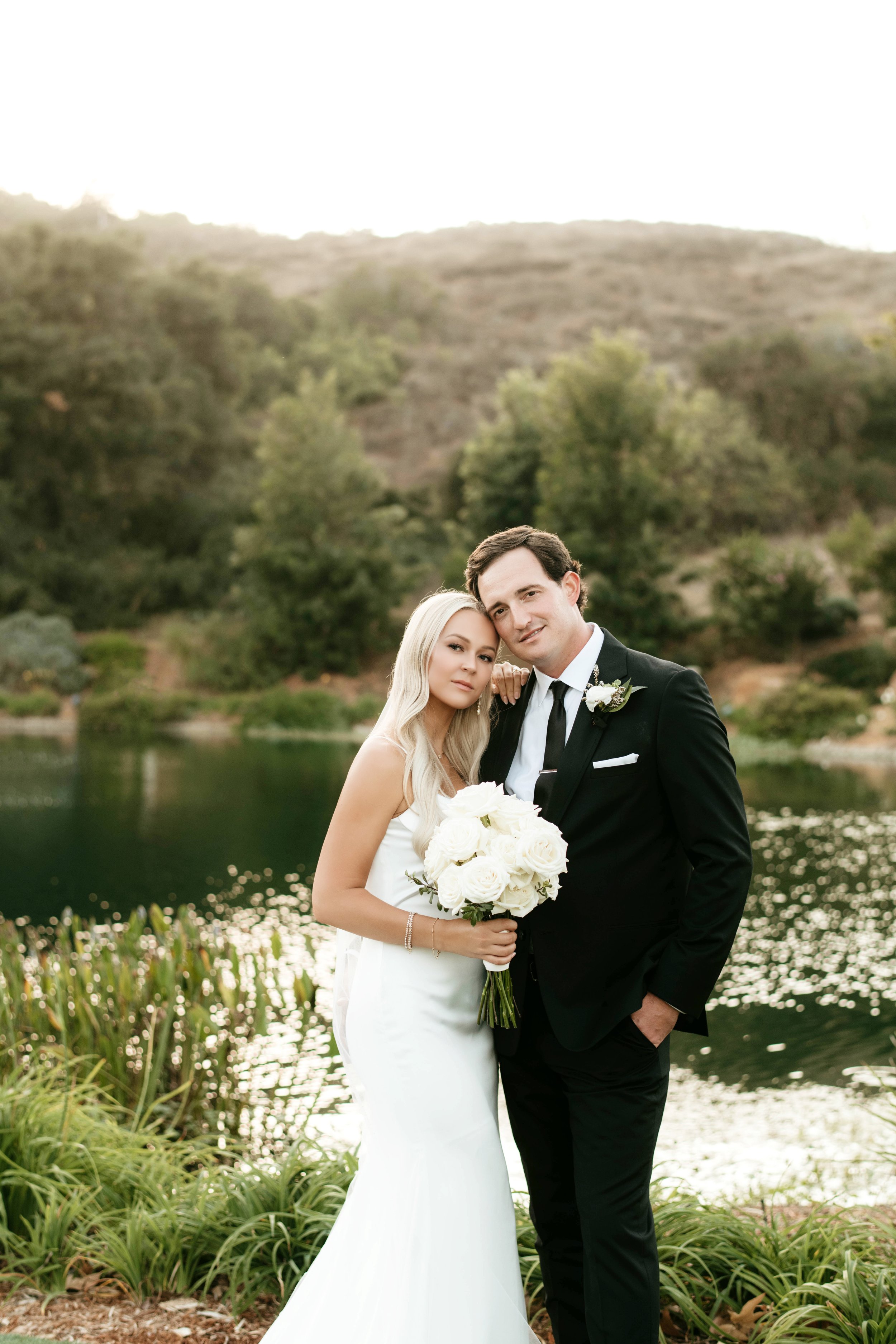 A bride and groom standing by a lake with hills and trees in the background. The bride has long blonde hair and wears a white wedding dress, holding a bouquet of white roses. The groom has dark hair, wears a black tuxedo, and is smiling. They are emb