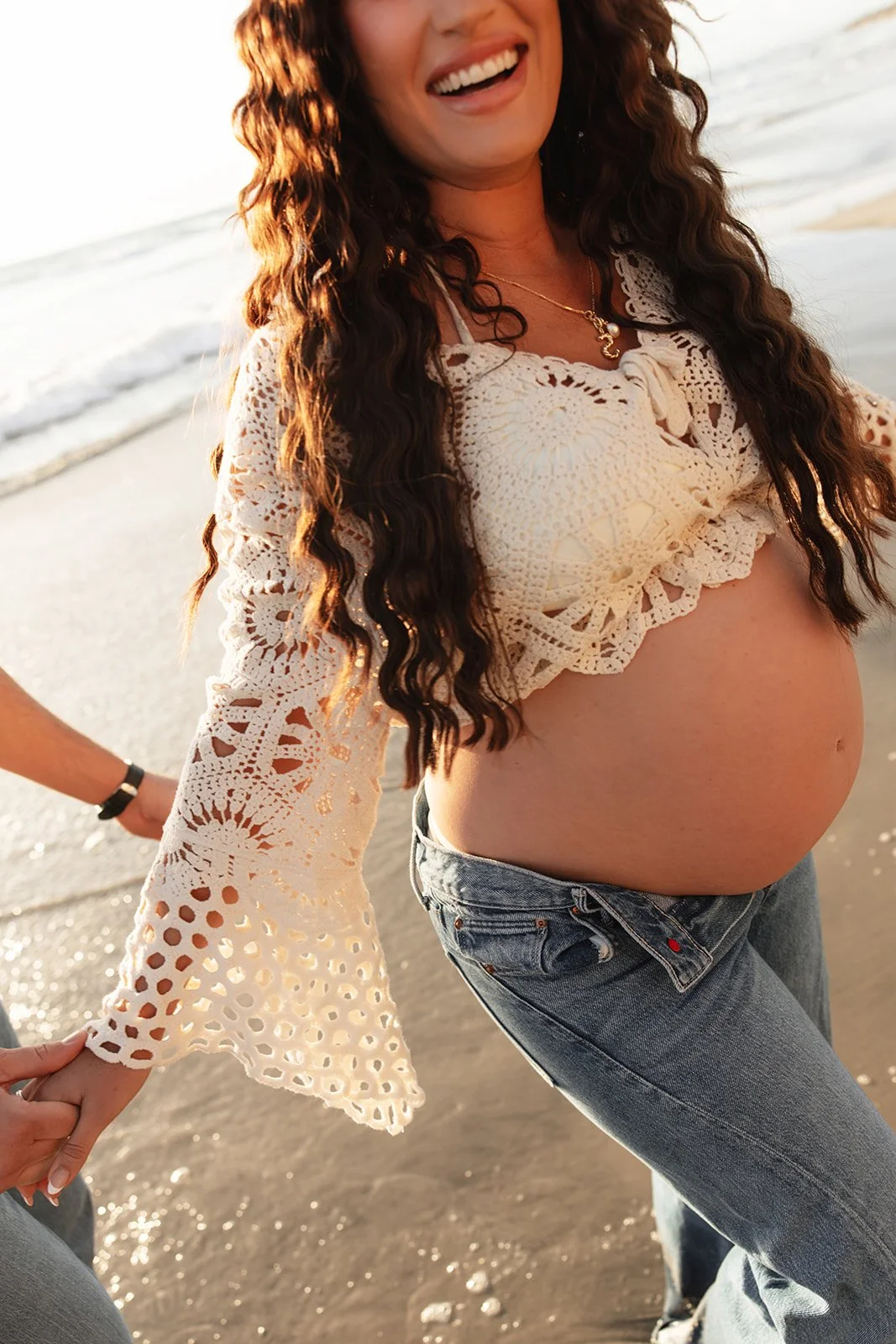 A pregnant woman smiling and holding hands on a beach during sunset, wearing a lacy cream top and jeans.