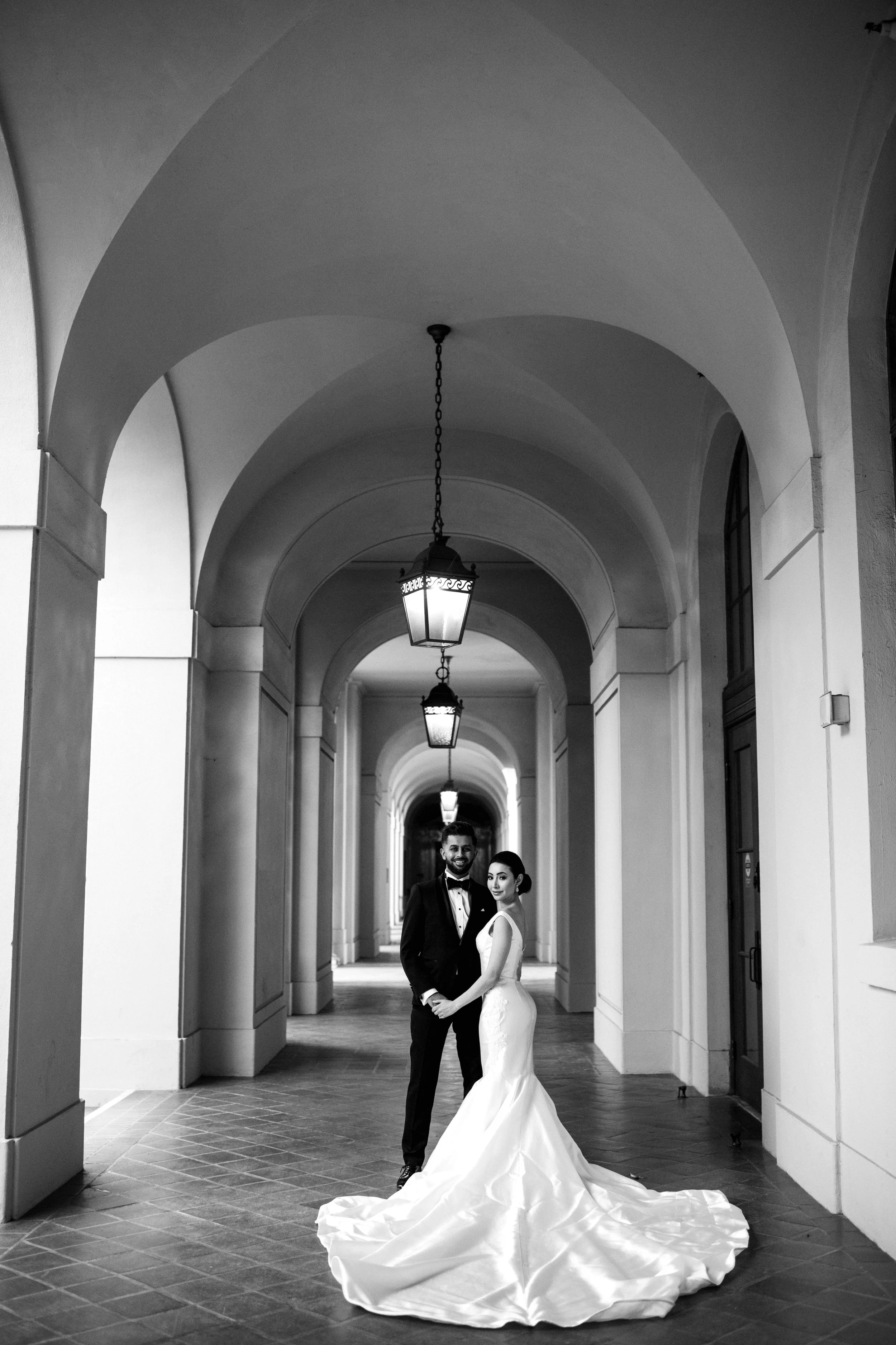 A bride and groom stand together in wedding attire in a covered walkway with arched ceiling and hanging lanterns, smiling at the camera.