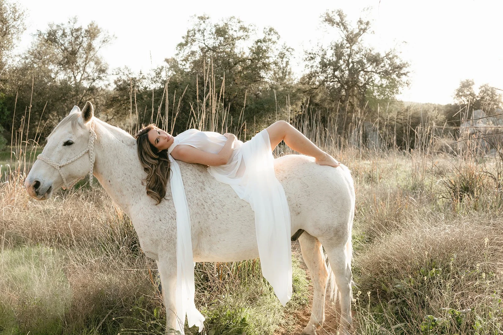 A woman in a white dress is lying on a white horse in a field at sunset, with trees in the background.