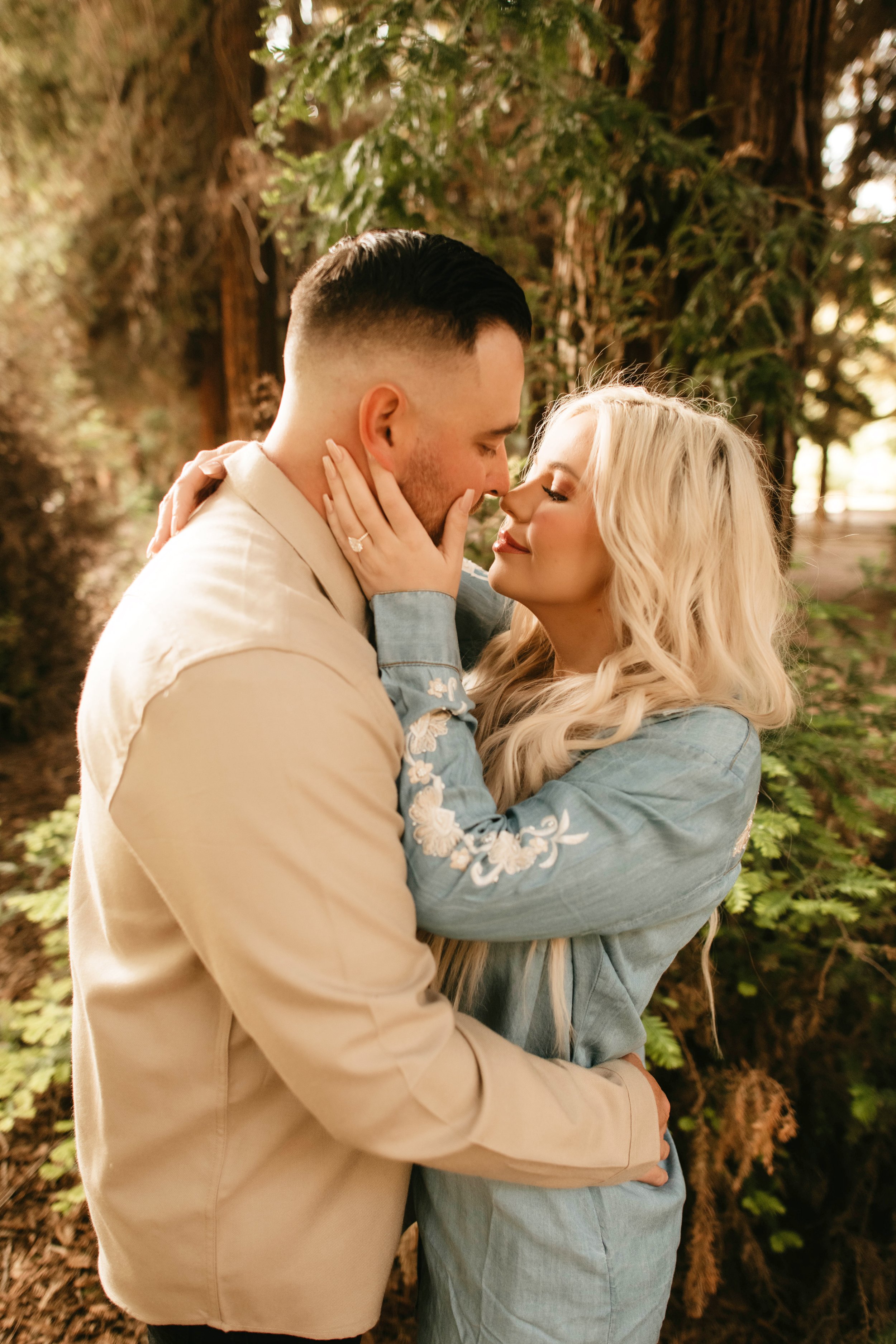 A couple shares an intimate moment outdoors among tall trees with green foliage, with the woman touching the man's face and both appearing happy and close.