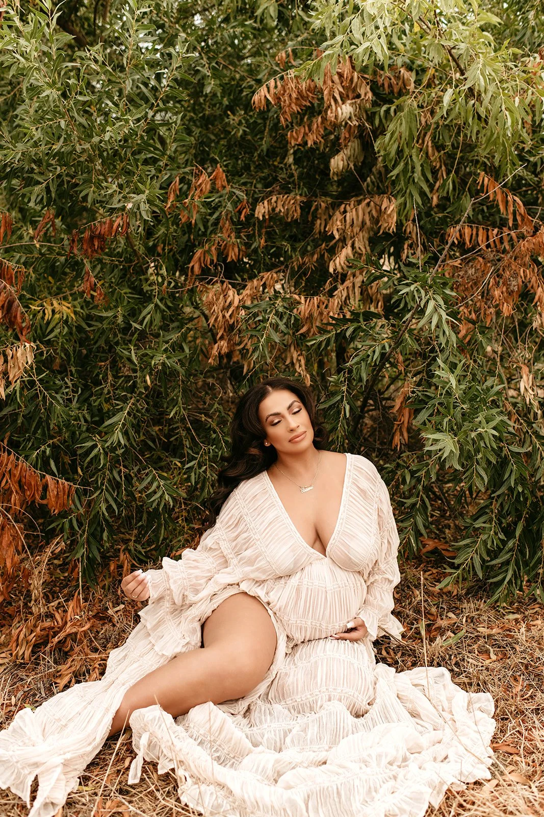 A woman with dark hair and makeup sitting outdoors on the ground among dried leaves and green bushes, wearing a cream-colored, flowing dress with a deep neckline.