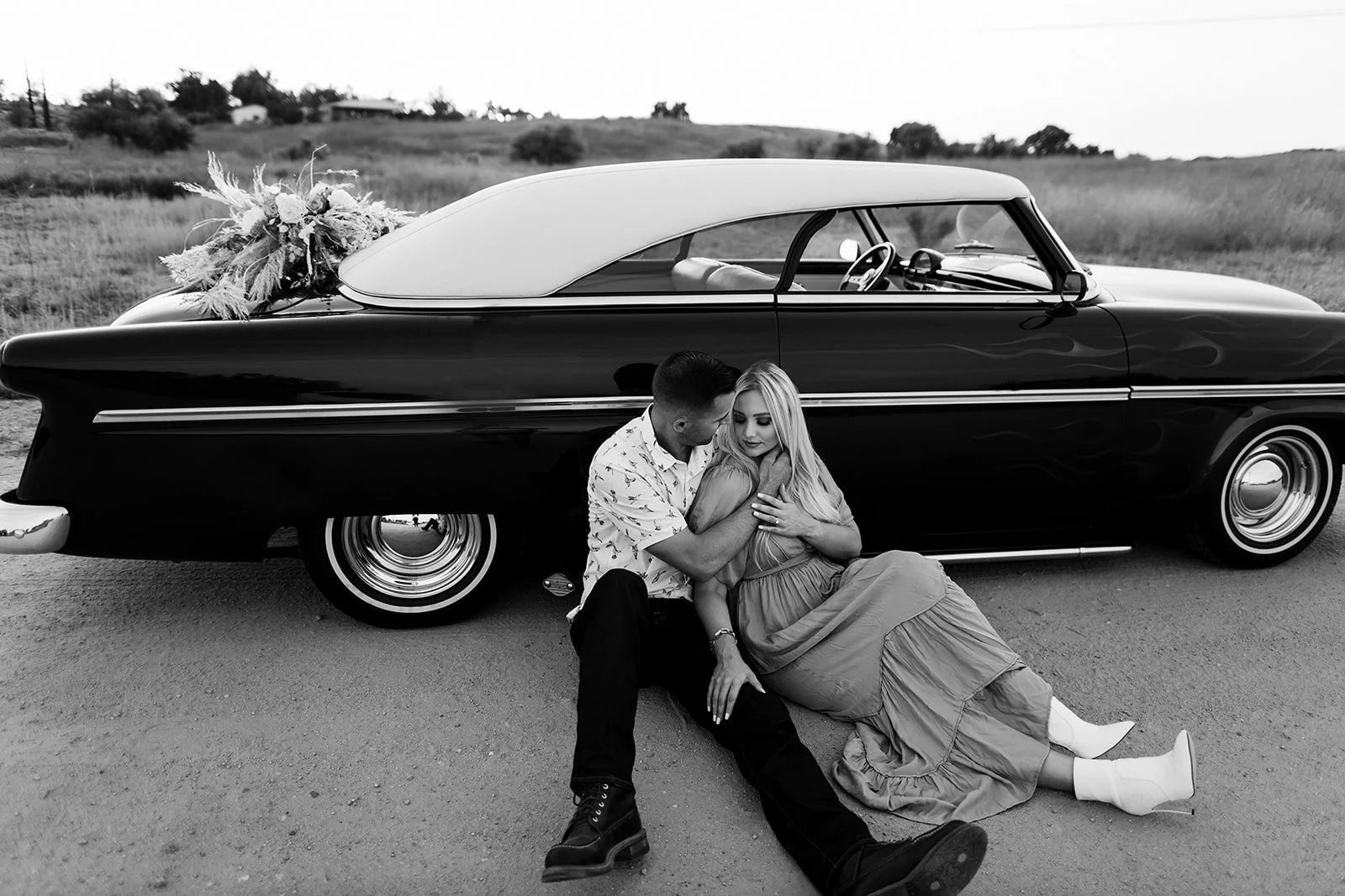 A black-and-white photograph of a couple sitting on the ground, embracing each other in front of a vintage car with a bouquet on top. The setting is outdoors with a hilly landscape in the background.