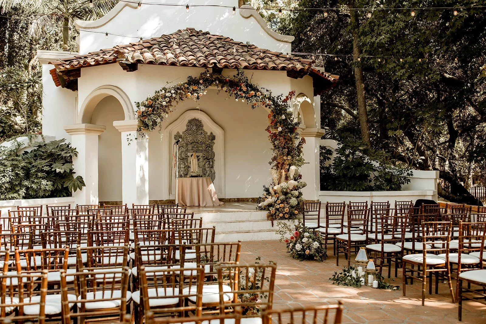 Outdoor wedding ceremony setup with rows of wooden chairs facing a small stage altar decorated with floral arrangements and a religious statue, under string lights and surrounded by trees.
