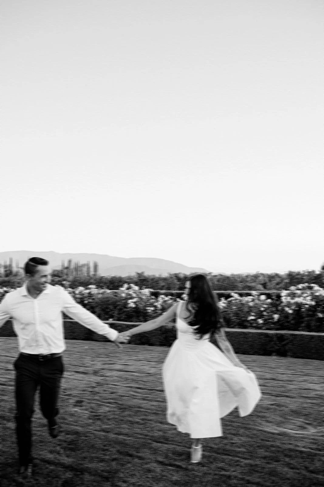 A black and white photo of a smiling man and woman holding hands while dancing outdoors in a field, with mountains in the background.