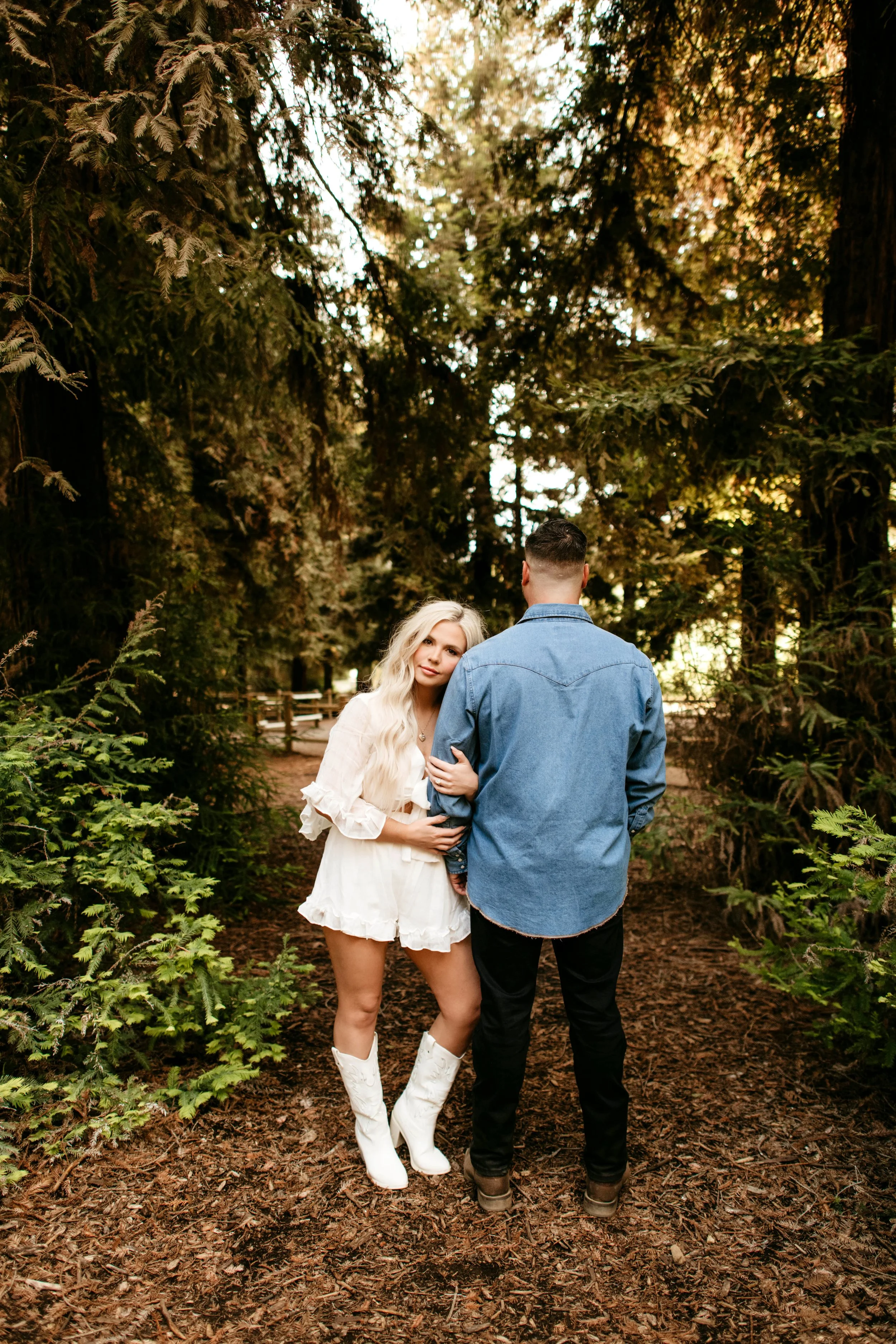 A woman with long blonde hair and a white dress stands next to a man with short hair and a denim shirt in a forested area with tall trees and a dirt path.
