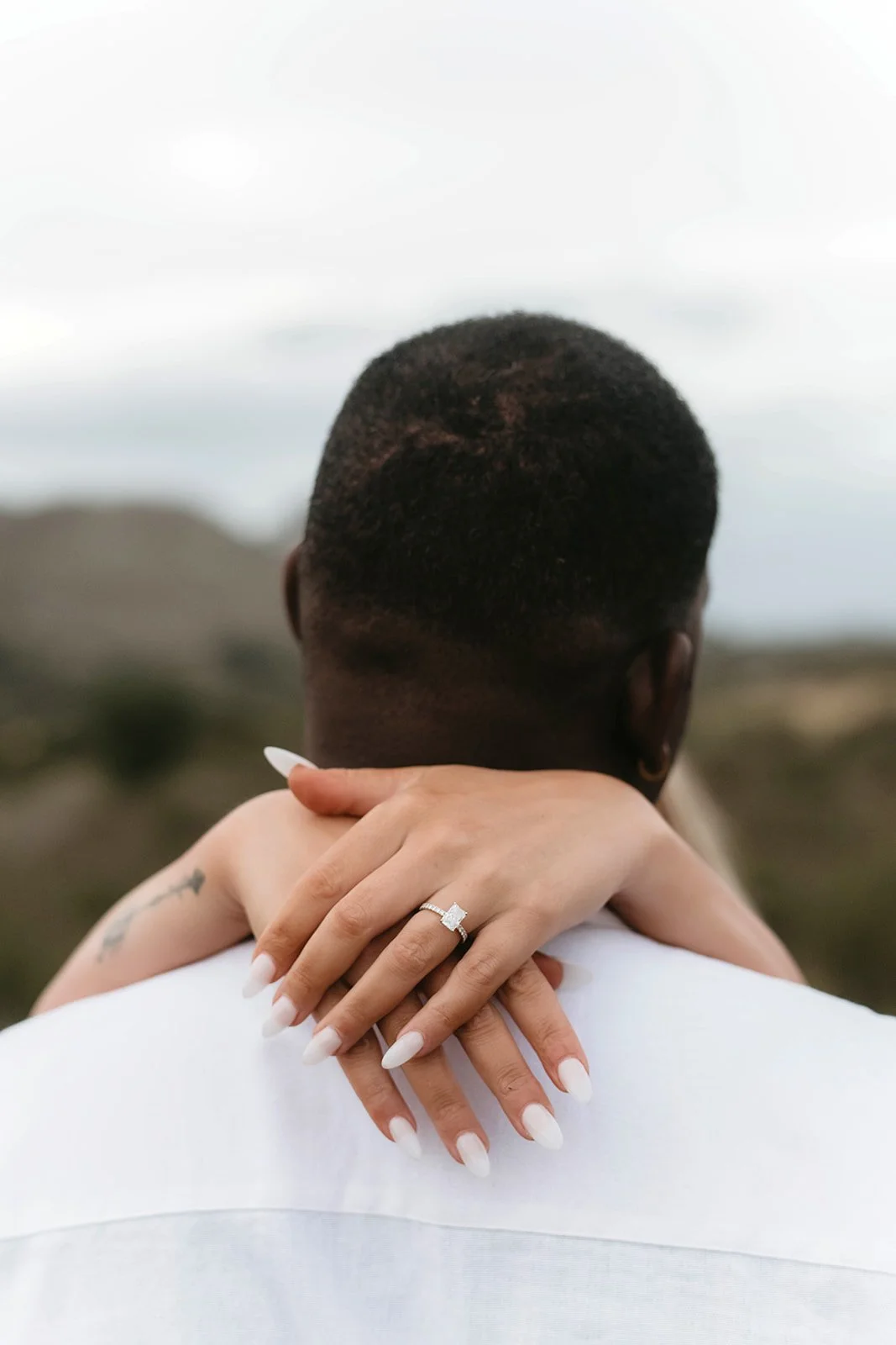 A couple embracing outdoors, viewed from behind, with a woman’s hands resting on a man’s shoulders, showing a diamond engagement ring.