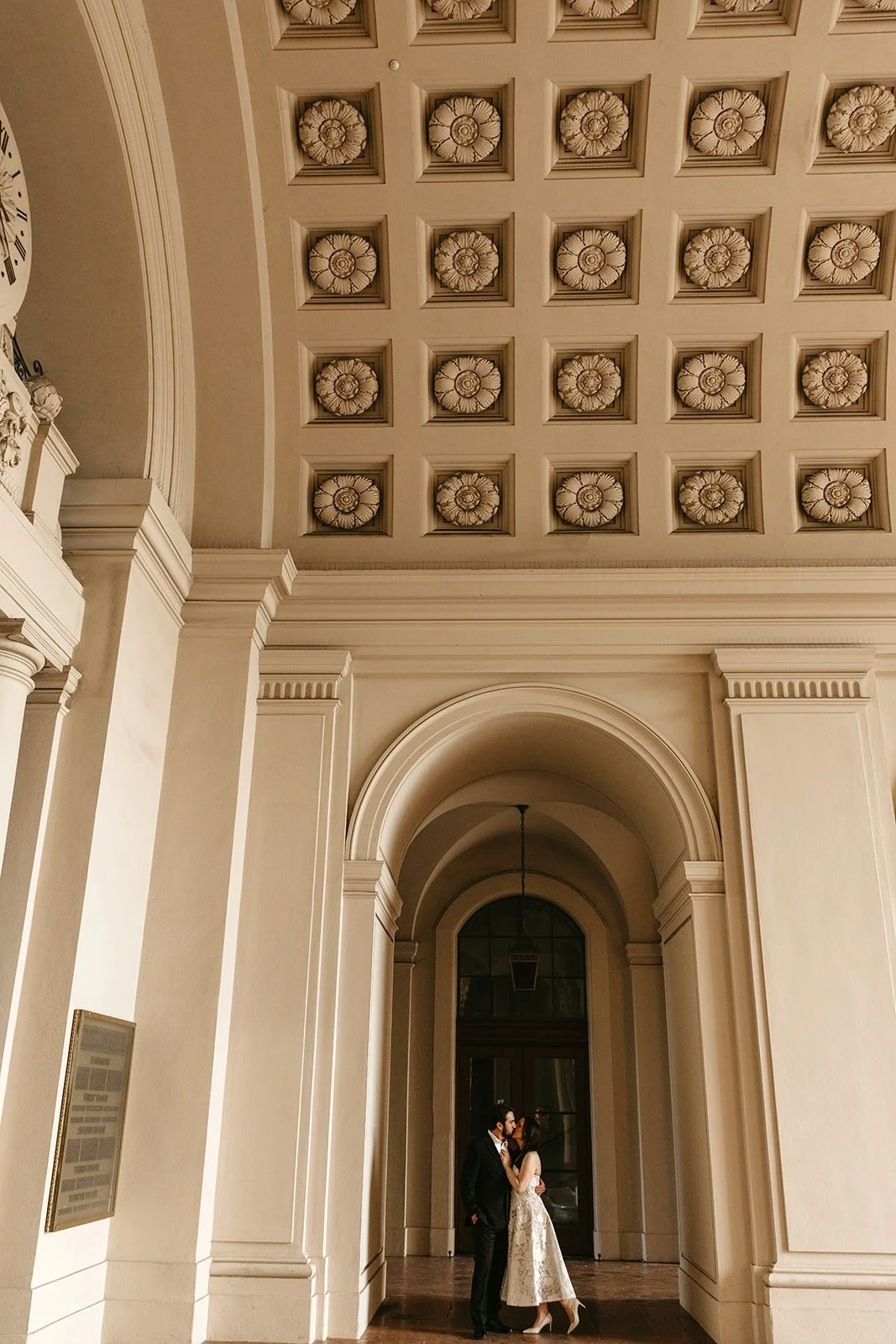 A couple dressed in wedding attire, a man in a black suit and a woman in a wedding dress, standing close together and kissing inside a grand, elegant hall with high ceilings, decorative columns, and detailed coffered ceiling.