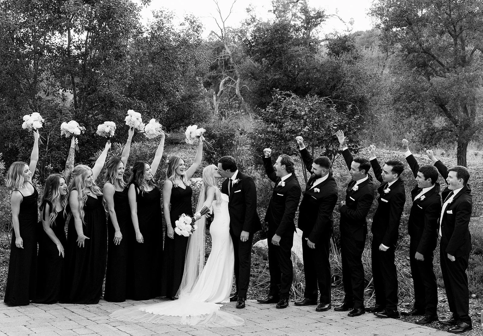 A black-and-white photo of a wedding party outdoors. The bride and groom are kissing in the center, surrounded by bridesmaids on one side and groomsmen on the other. The bridesmaids are holding bouquets and the groomsmen have boutonnieres. The guests