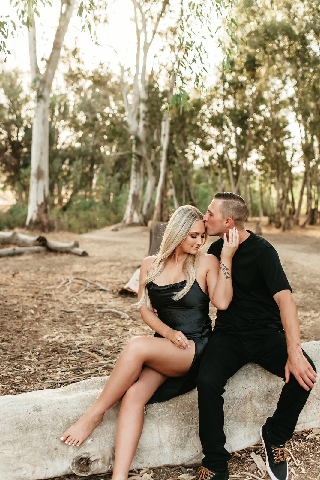 A couple sitting on a fallen log in a wooded area, with the man kissing the woman's forehead. They are dressed in black, and the woman has long blonde hair. The background features trees with sunlight filtering through.