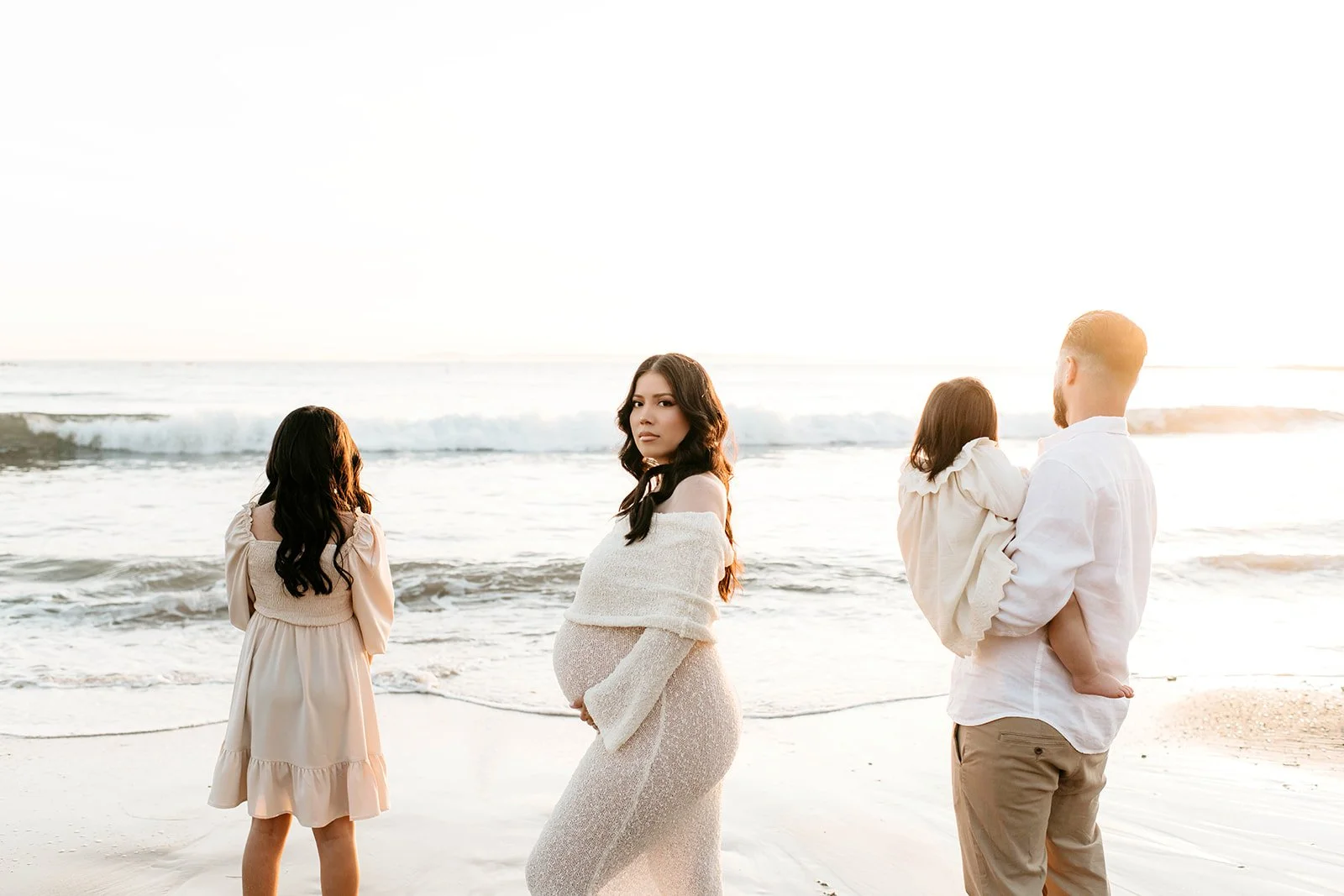 A pregnant woman standing on the beach with four women, one man holding a child, near the ocean at sunset.