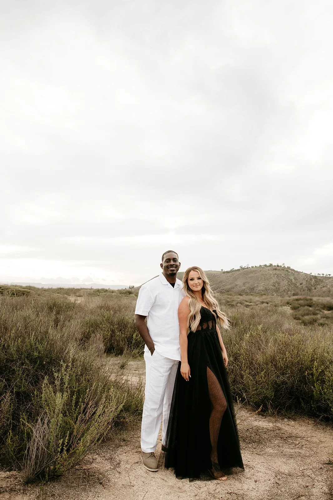 A couple standing outdoors in a grassy field with hills in the background, smiling at the camera. The man is wearing a white shirt and pants, and the woman is wearing a black dress.
