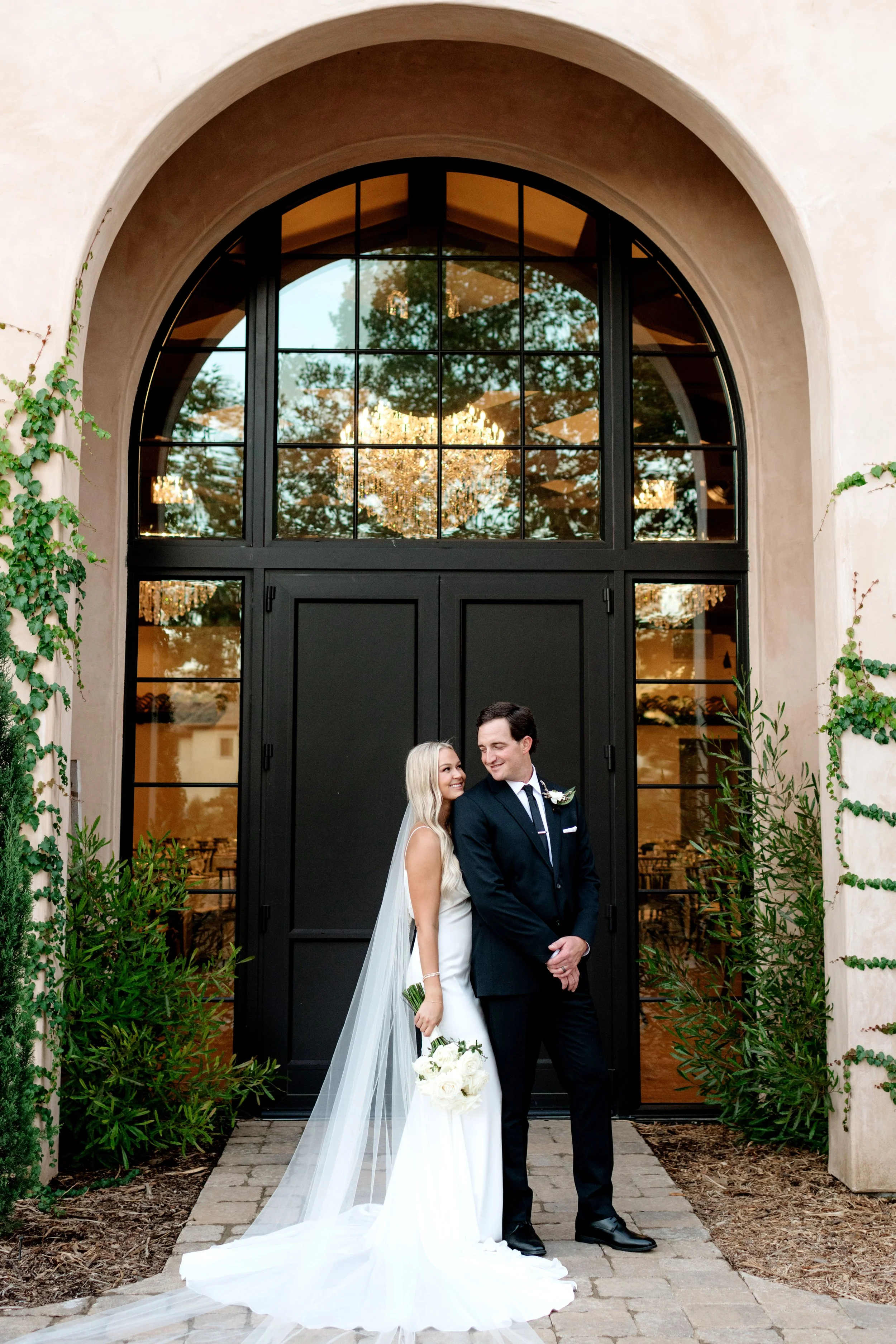 A bride and groom standing outside a building with large black arched windows, smiling at each other. The bride is in a white wedding dress with a veil, holding a bouquet of white roses. The groom is in a dark suit with a boutonniere. There are green plants on either side of them, and the building has ivy growing on the walls. Reflections of a chandelier and trees are visible in the windows.