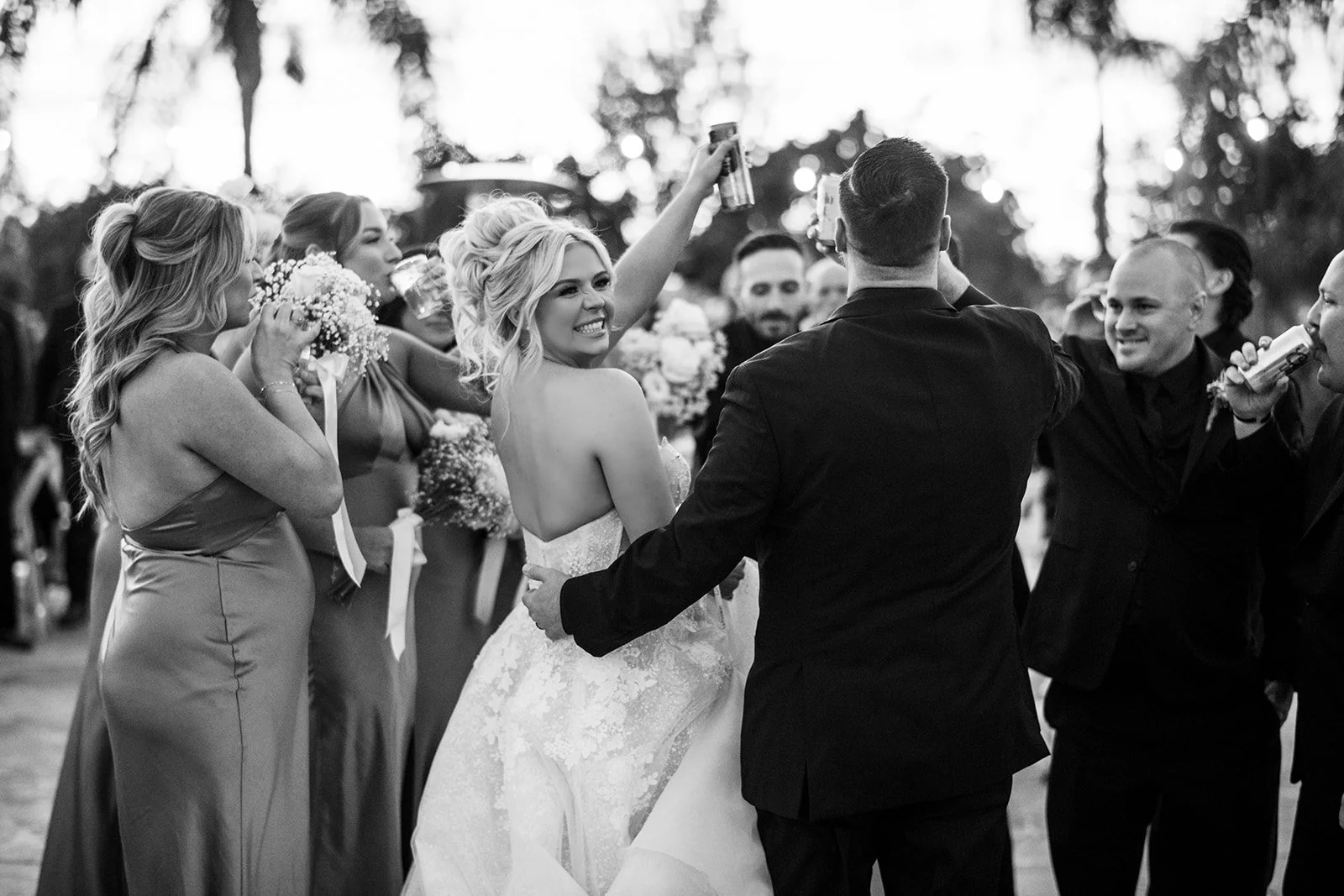 A bride turns to smile at a man in a tuxedo during a celebration, surrounded by women in formal dresses and men, with some holding drinks, outdoors with trees in the background.