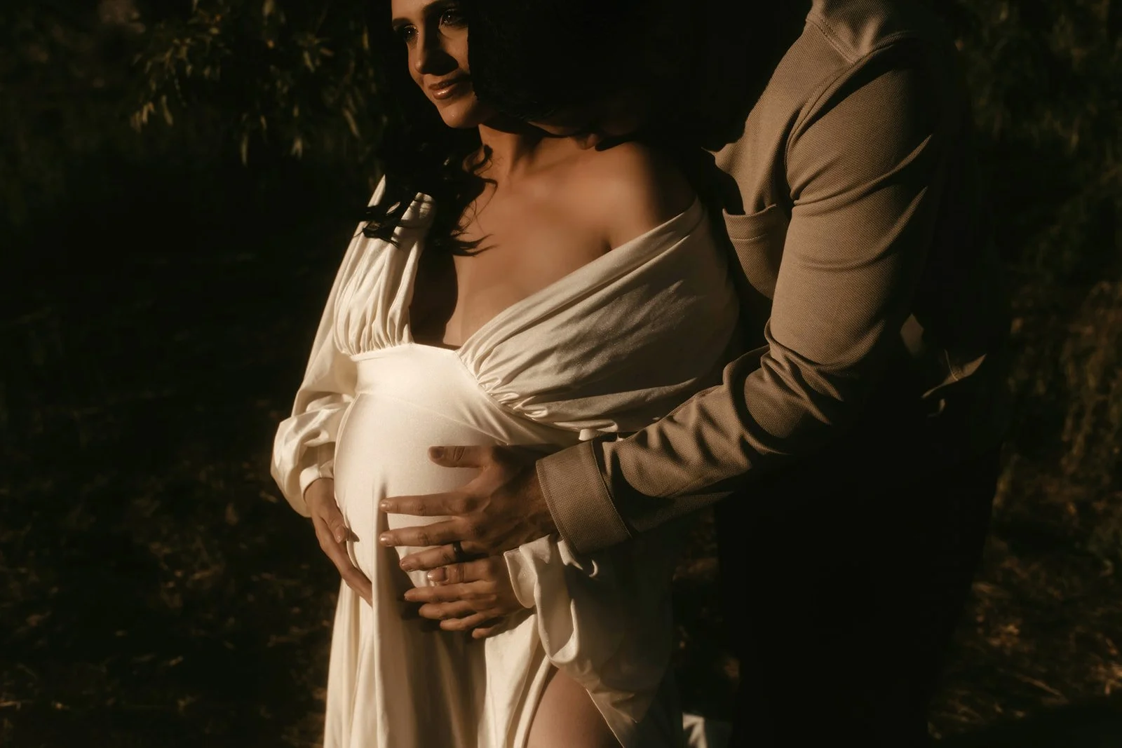 A pregnant woman with dark hair in a white dress holding her belly, with a man behind her holding her belly with both hands, both in warm lighting outdoors.