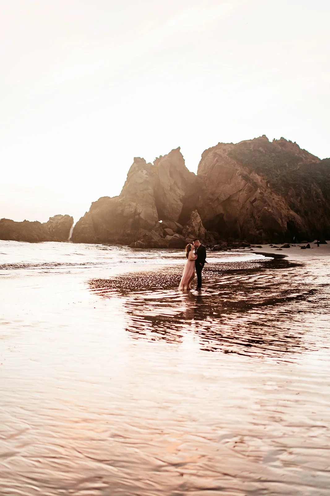 A couple in formal attire embracing in shallow water on a beach with large rock formations and cliffs in the background during sunset.