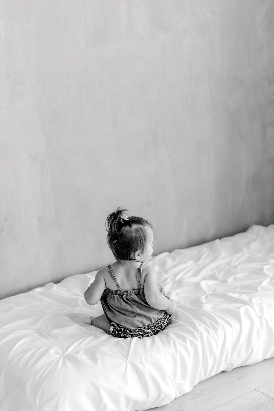 A young girl with a ponytail sitting on a bed with white bedding, facing away from the camera in a minimalistic room.