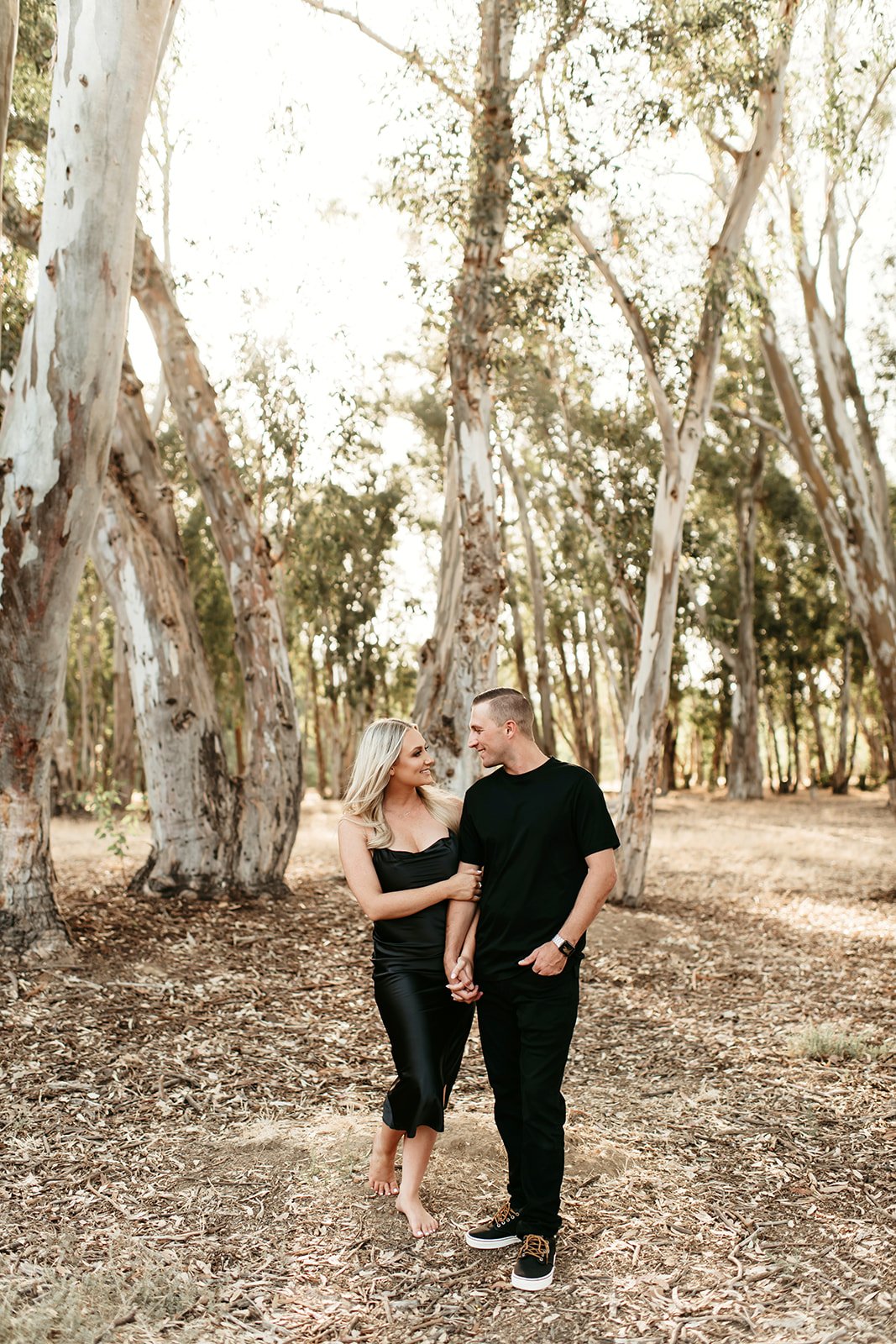 A couple walks hand in hand through a wooded area with large trees, sunlight filtering through the leaves, and dry leaves on the ground.