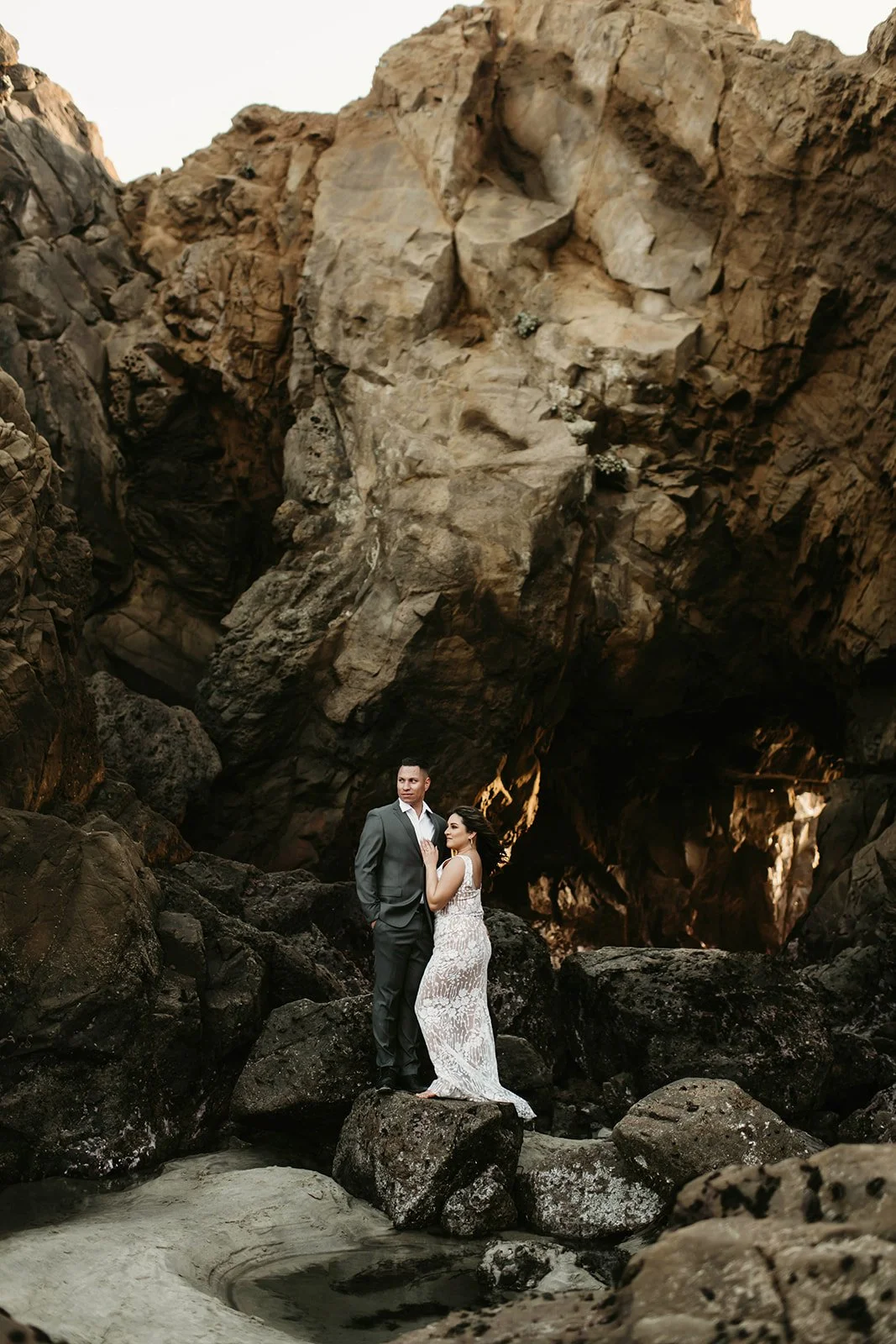 A couple dressed in formal attire on a rocky beach, with rugged cliffs in the background and a dark cave opening behind them.