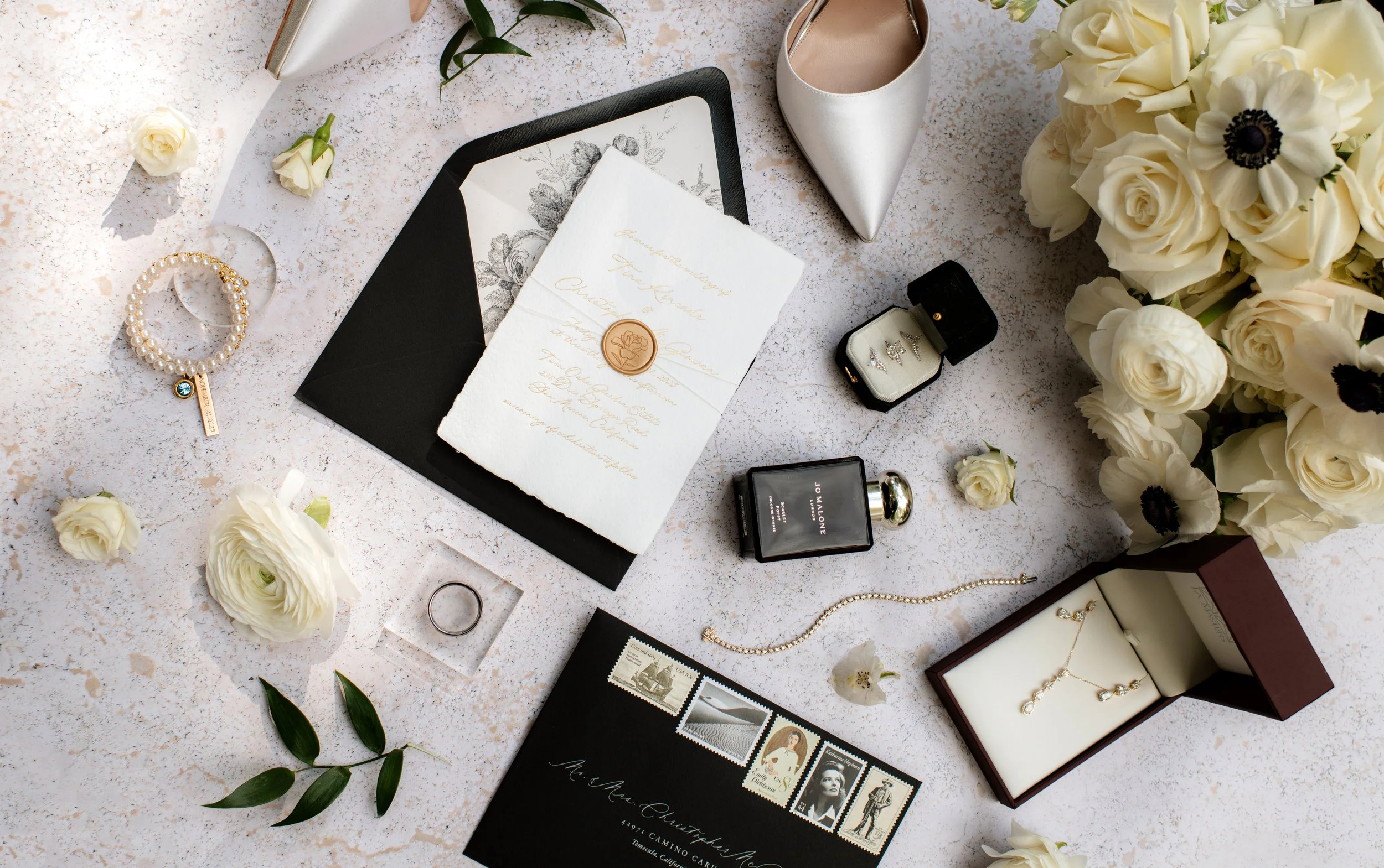 A flat lay of wedding items including a wedding invitation with gold lettering, white roses, a pearl bracelet, jewelry, a wedding ring, a box with earrings, a perfume bottle, and wedding shoes, all arranged on a light surface with scattered flowers a