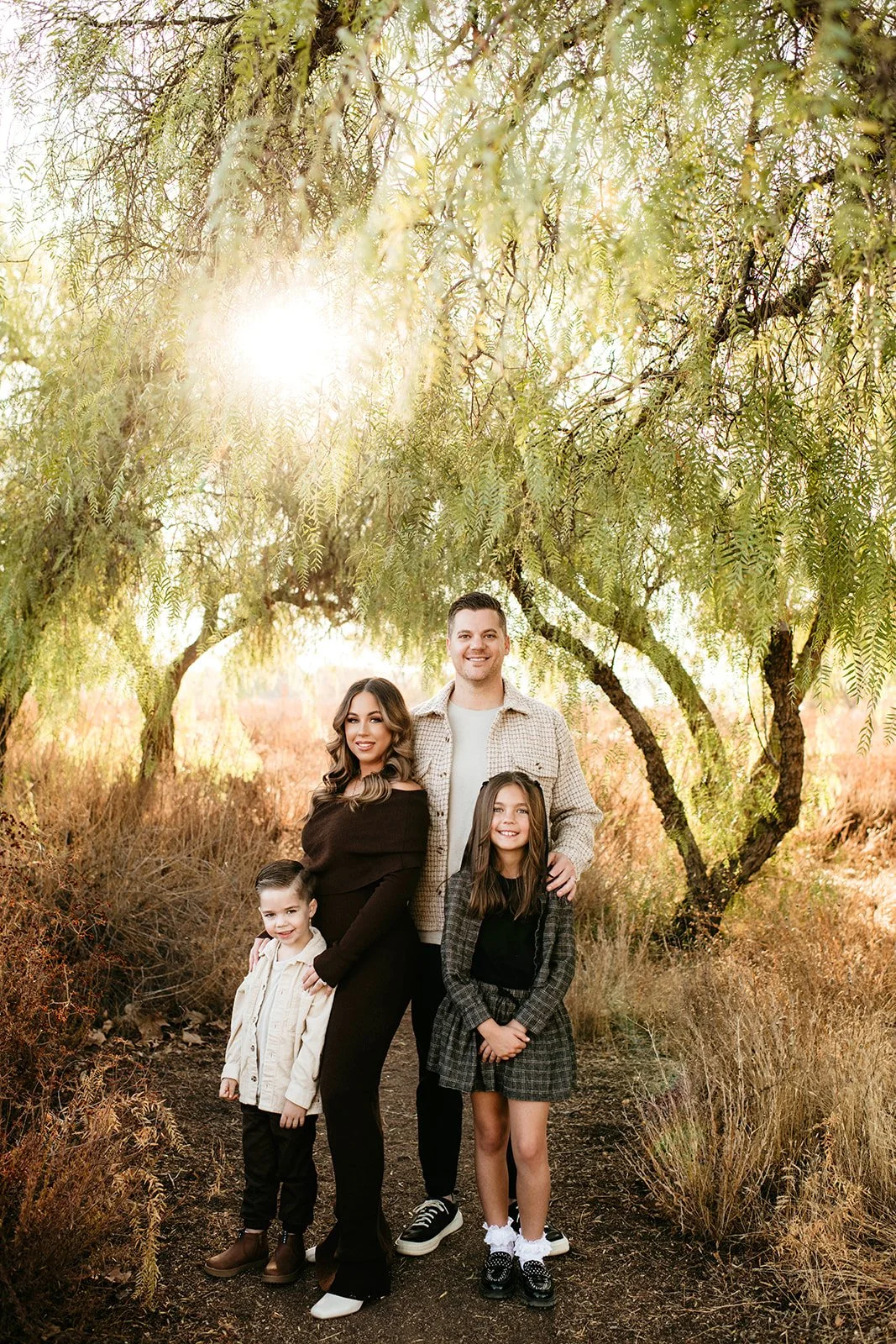 A family of four stands on a wooded trail with trees and sunlight in the background. The adults and children are smiling and dressed in casual, stylish clothing.