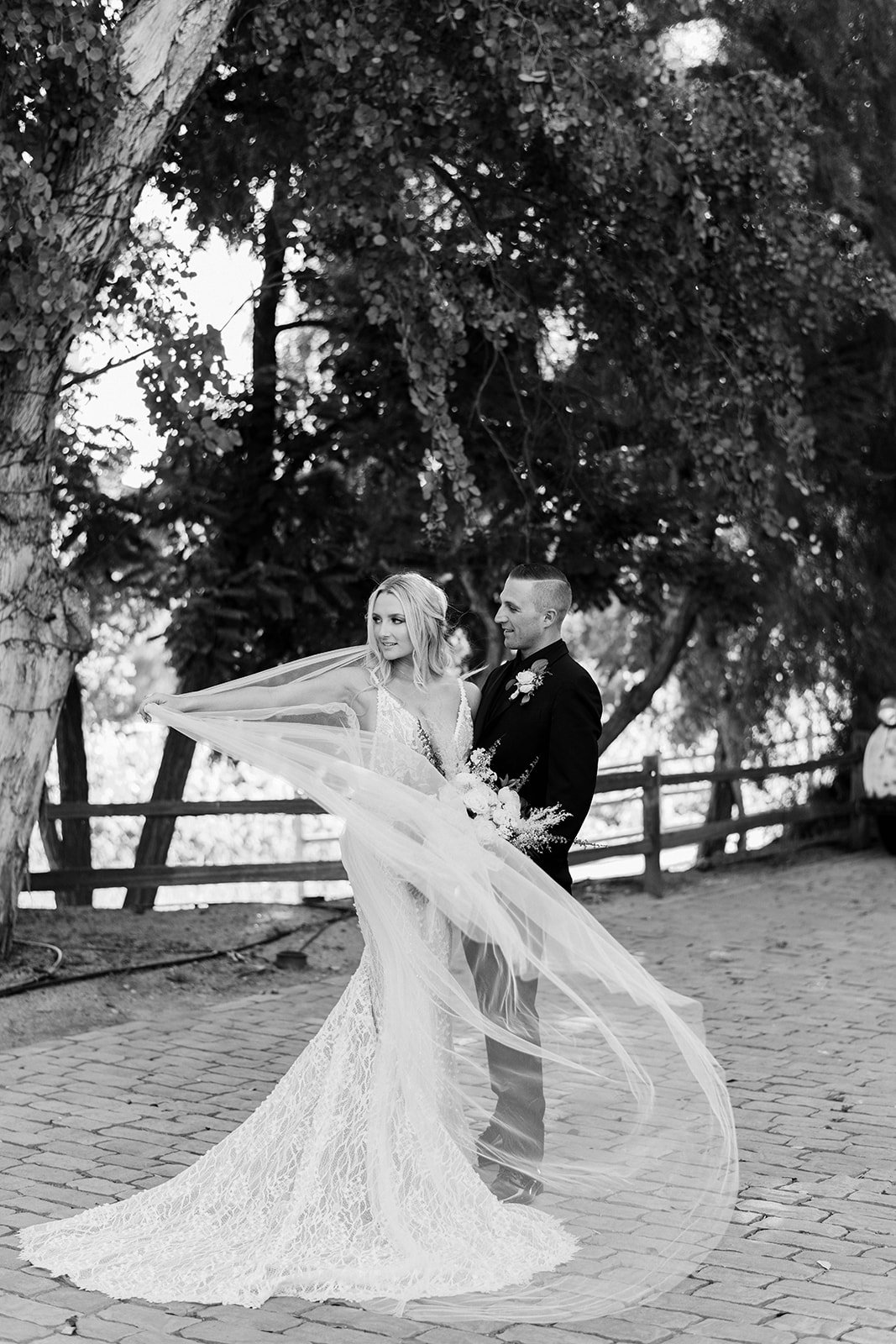 Black and white photo of a bride and groom outdoors, the bride wearing a long lace wedding dress with a veil, and the groom in a suit with a boutonnière, standing on a cobblestone path beneath large trees.