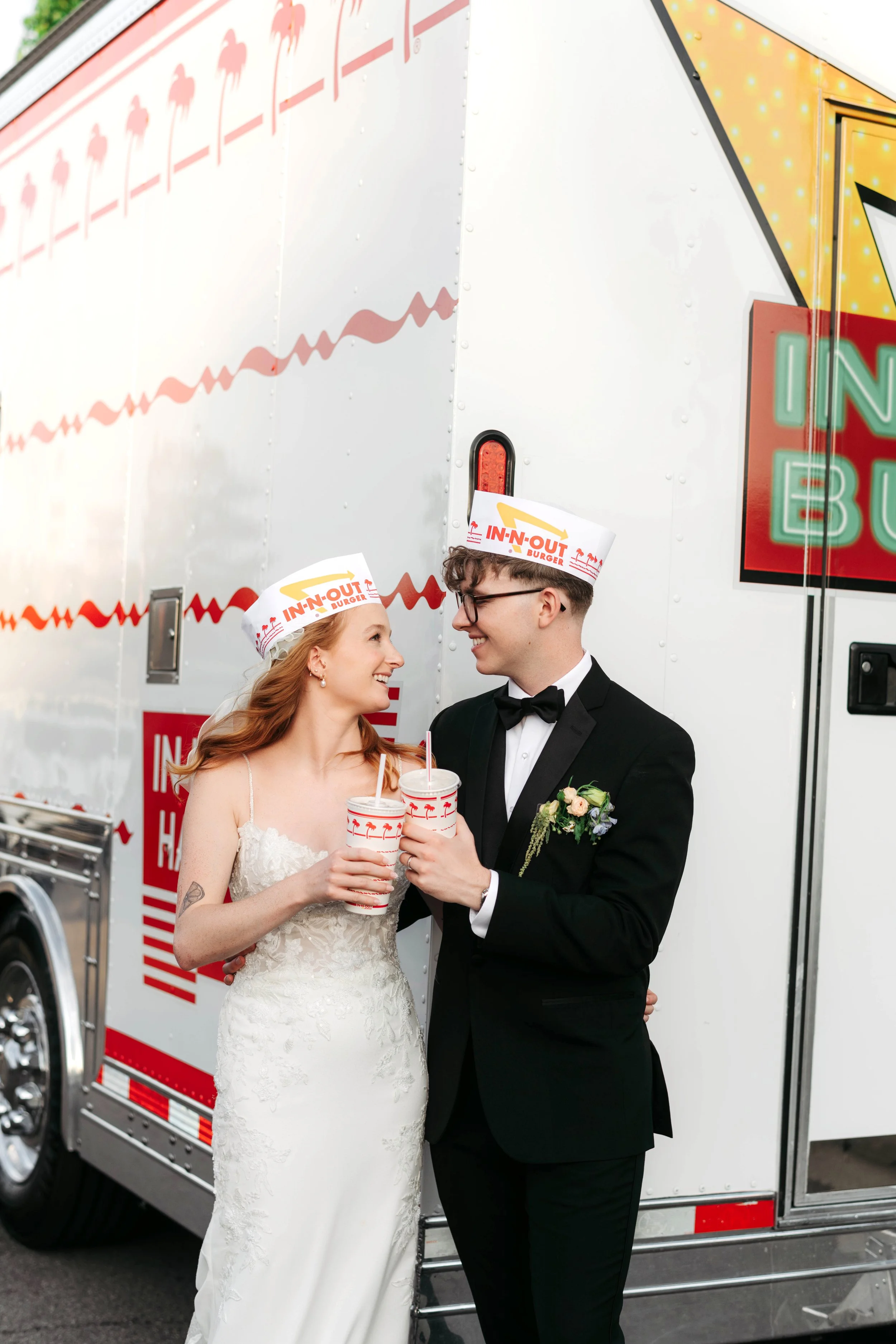 A newlywed couple stands in front of an In-N-Out Burger food truck, smiling and holding drinks, during their wedding celebration.