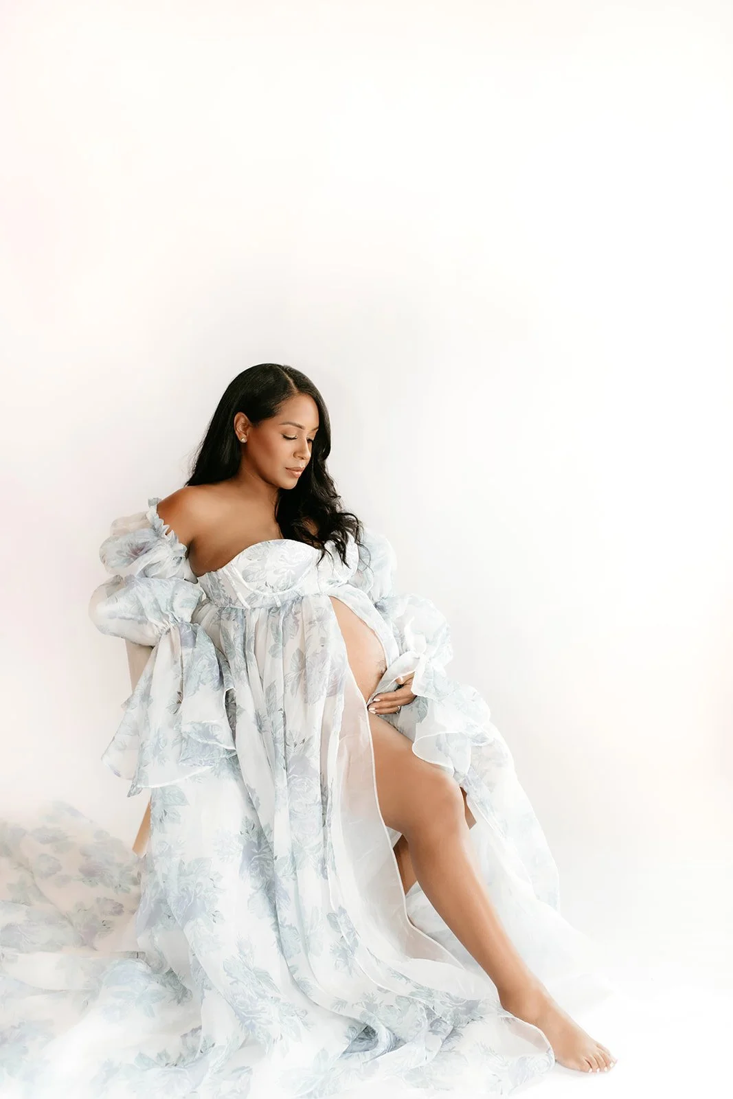 Woman in a white floral gown sitting against a white wall, gazing down, with her hand on her belly and one leg extended.