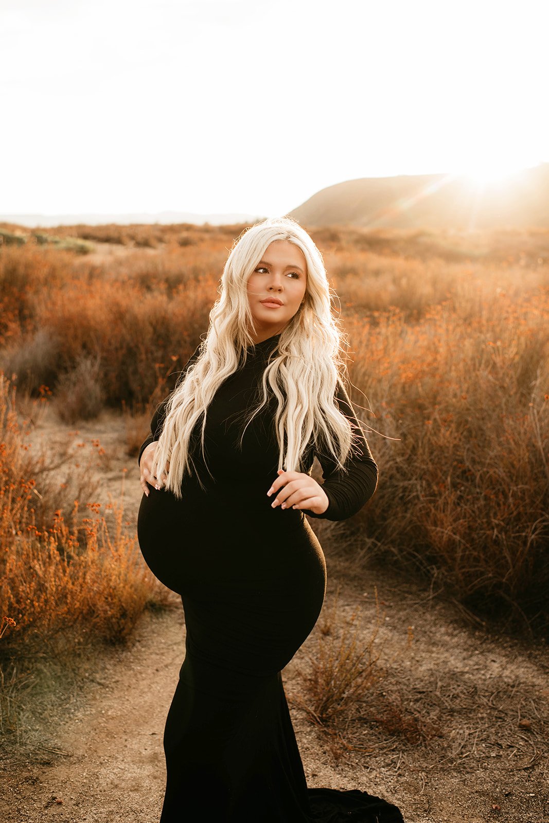 A pregnant woman with long blonde hair standing outdoors during sunset in a landscape with dry bushes and hills, wearing a form-fitting black dress.