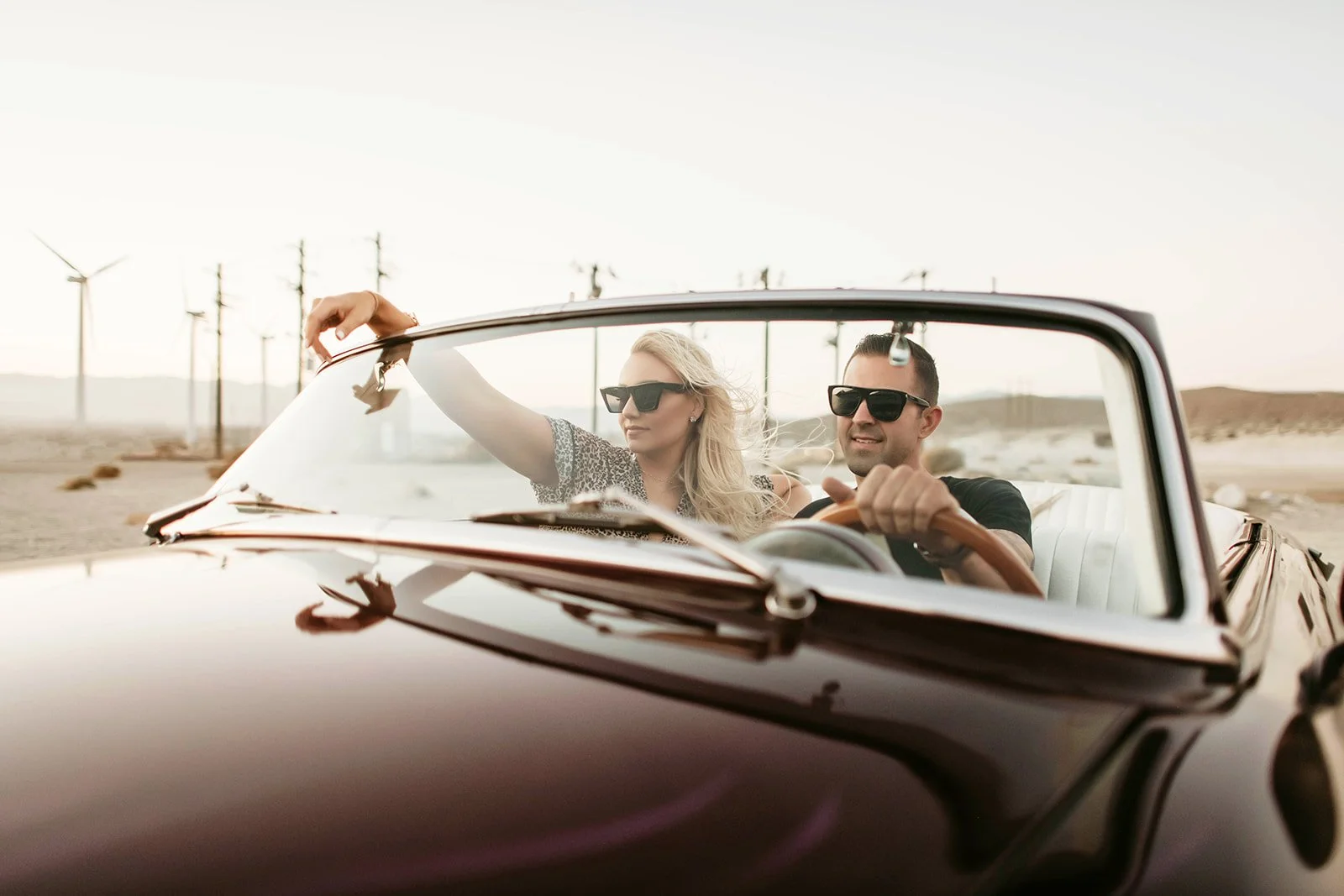 A man and woman wearing sunglasses driving a black convertible car in a desert area with power lines in the background.