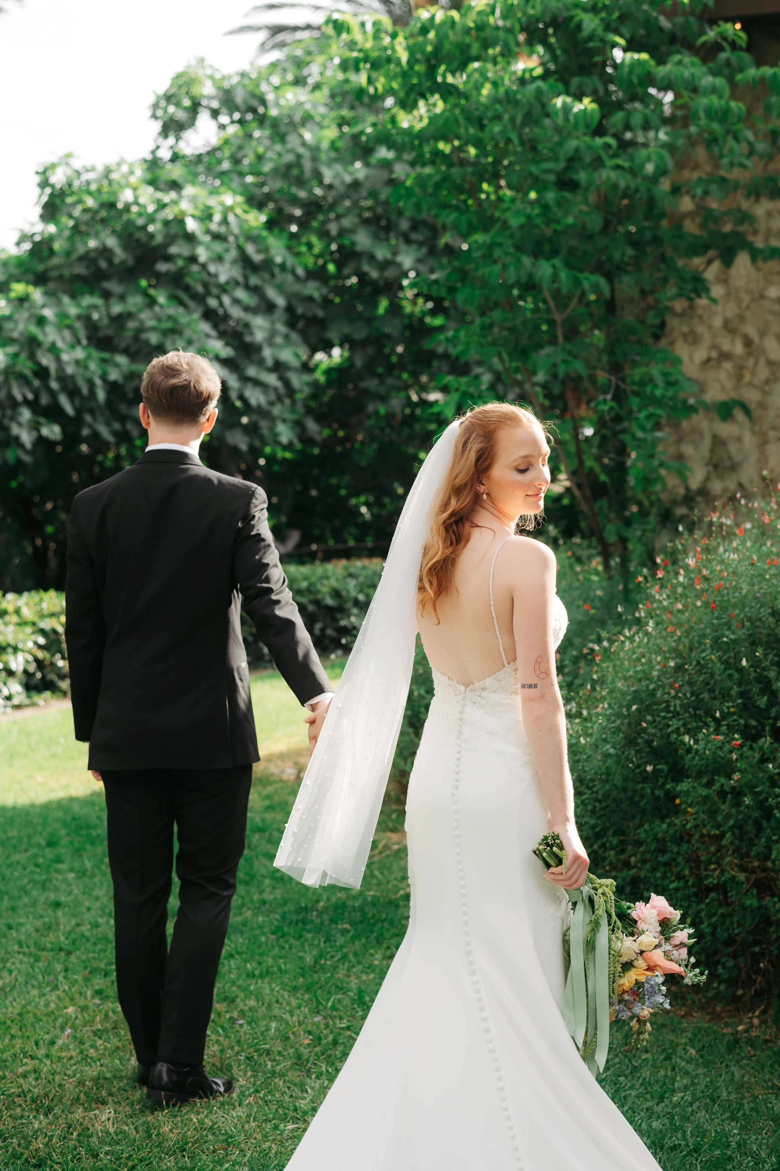 A bride and groom holding hands outdoors, with greenery and trees in the background. The bride has long red hair and is wearing a white wedding gown and veil, while the groom is dressed in a black suit with his back turned to the camera.