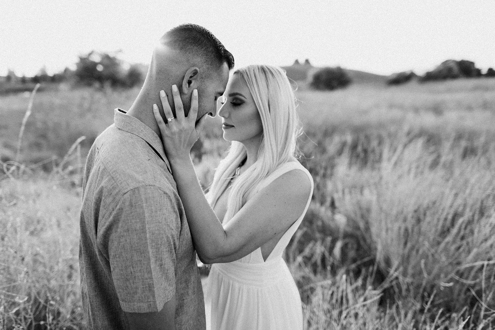 A black-and-white photo of a man and woman in an outdoor field, holding each other closely with their foreheads touching. The woman is wearing a sleeveless dress and has long hair, while the man is wearing a collared shirt and has short hair. The sce