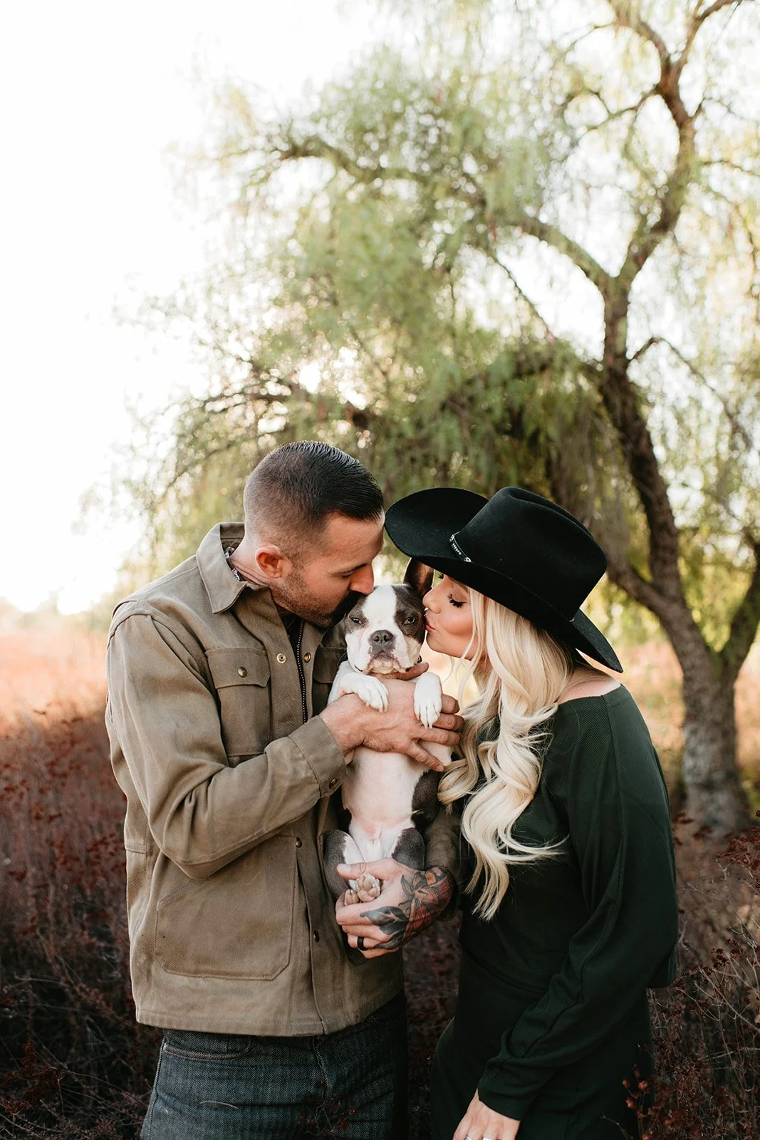A couple holding a puppy in an outdoor setting with trees in the background. The man is kissing the puppy's head, and the woman is giving a gentle kiss to the puppy as well. The woman is wearing a large black hat and has long blonde hair, while the m