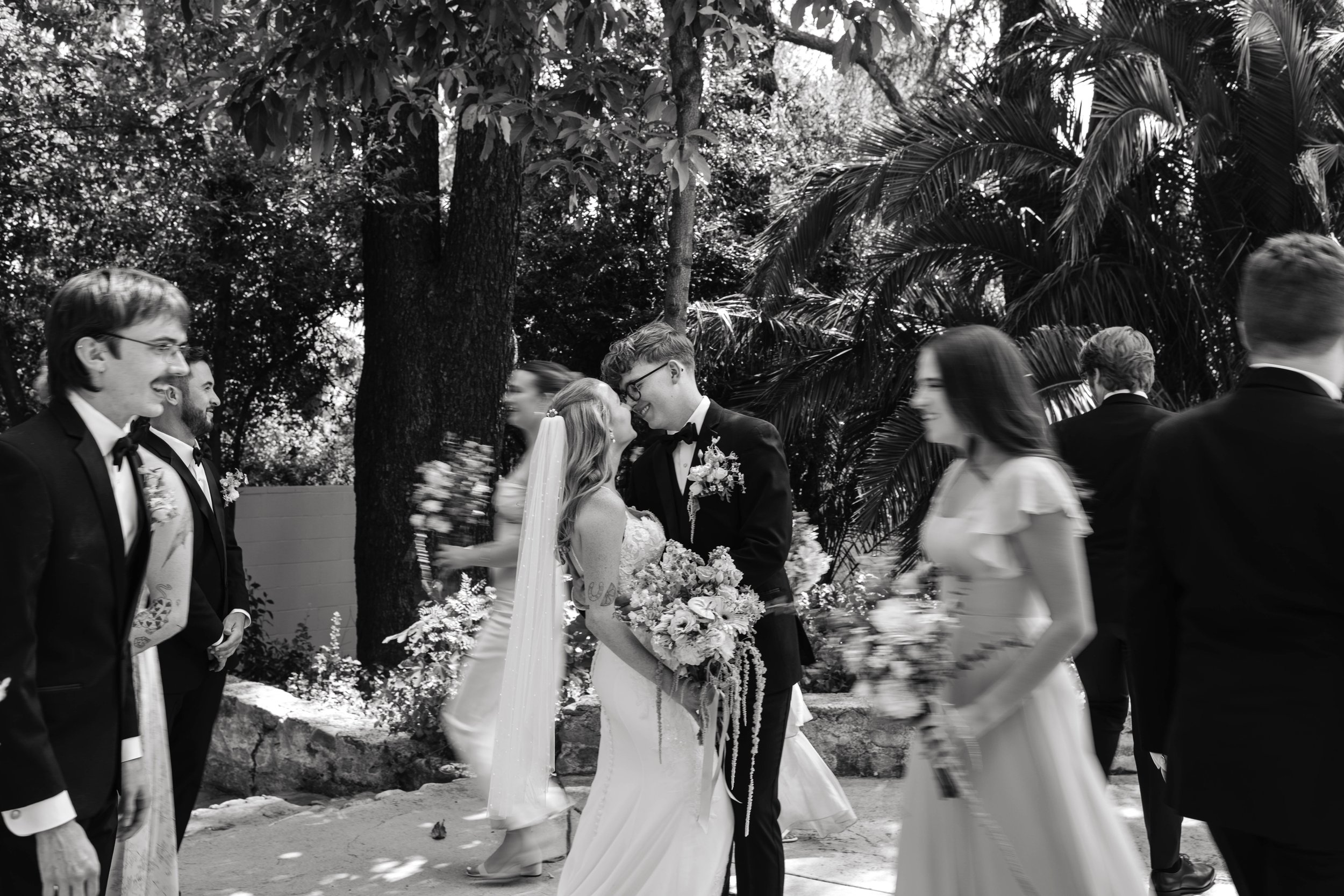 A wedding ceremony outdoors with a bride and groom kissing, surrounded by wedding guests in formal attire, under tall trees and lush greenery.
