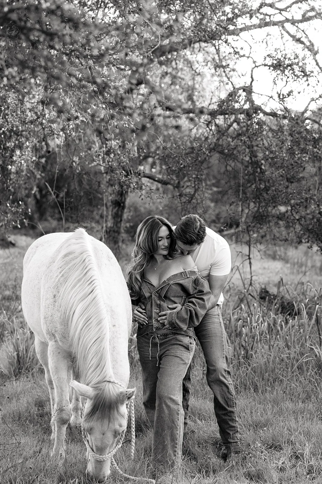 A black-and-white photo of a couple standing in a grassy field, with a white horse nearby. The man is leaning in and touching the woman's arm, while the woman looks down with a gentle expression. There are trees and a clear sky in the background.