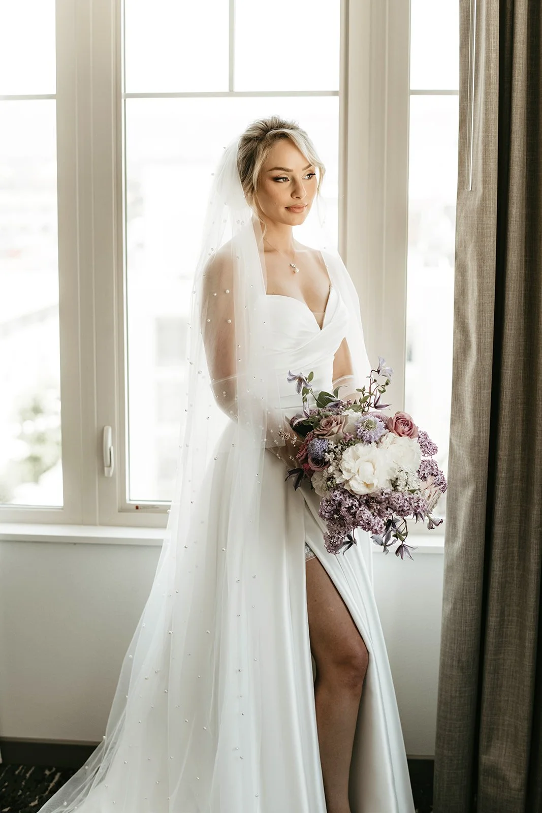 A bride in a white wedding gown holding a bouquet of flowers, standing by a window with natural light, looking thoughtfully to the side.