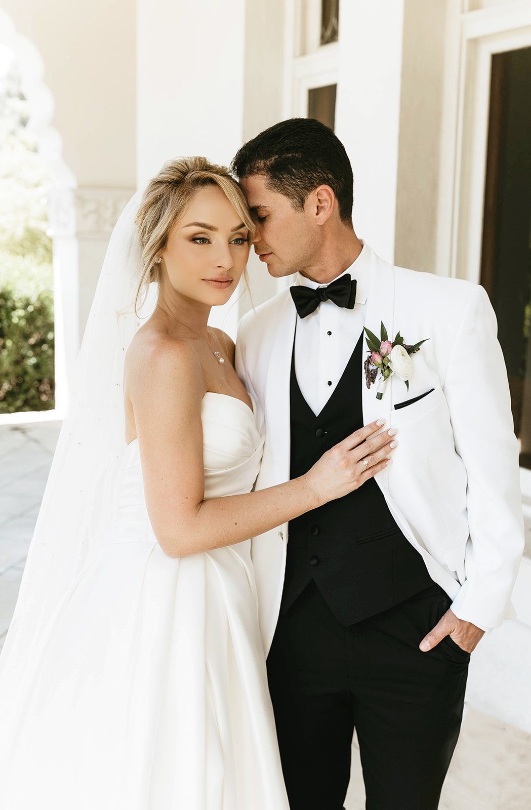 A bride and groom in wedding attire, standing close together with their foreheads touching, in a bright outdoor setting.