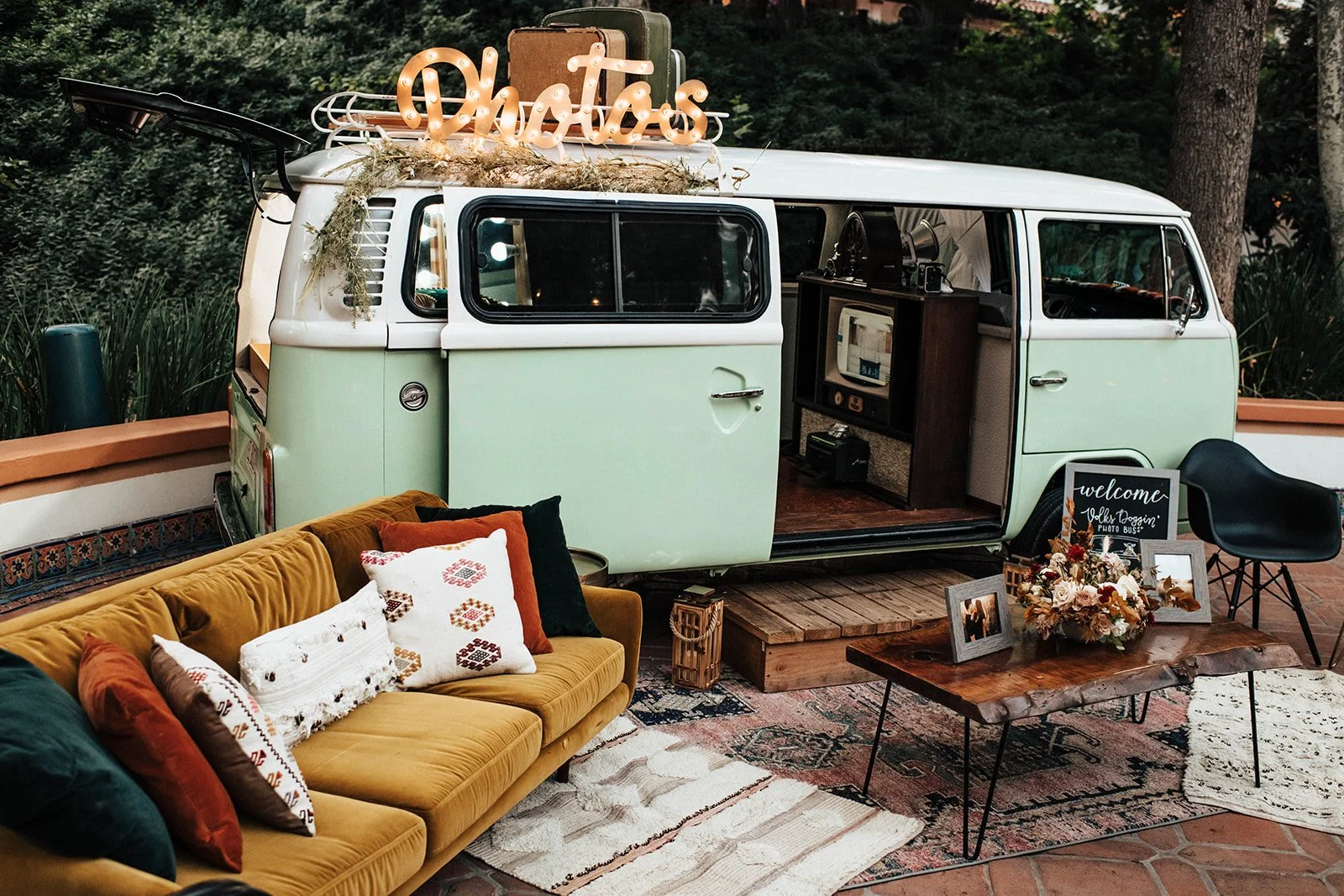 Decorated vintage green and white camper van set up as a cozy outdoor lounge with sofa, pillows, coffee table with flowers, rustic wood floor, and signs welcoming guests, surrounded by trees.