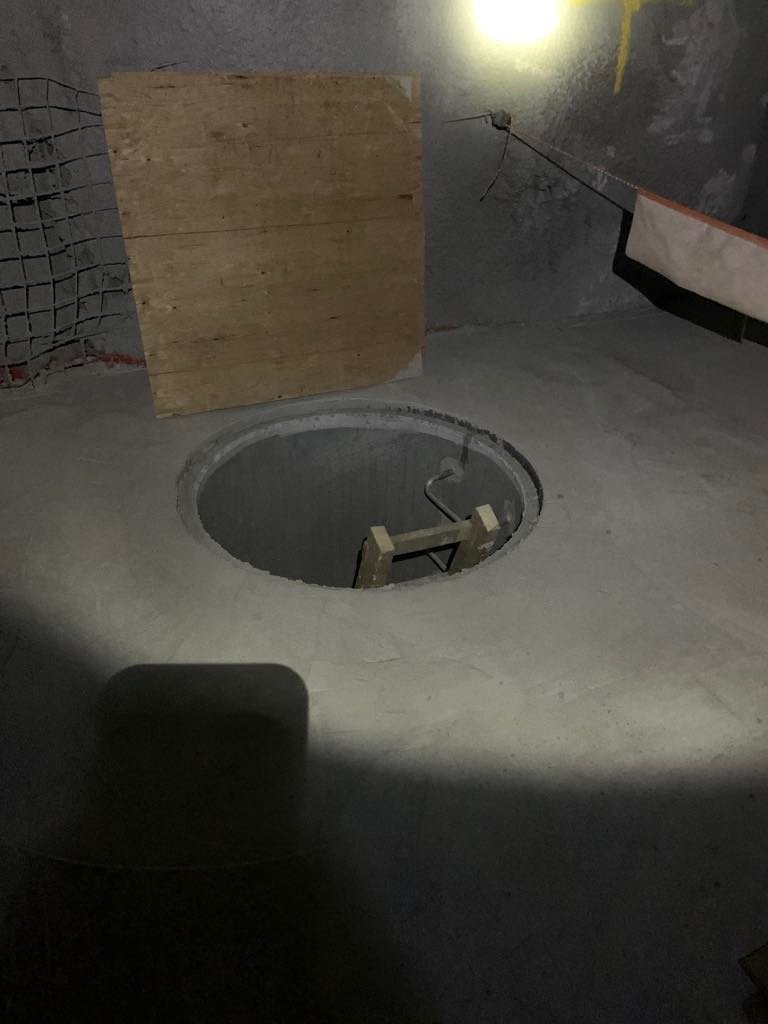 An unfinished mining space with a circular hole in the concrete floor, surrounded by a metal frame with a ladder inside, and a wooden board leaning against the wall.