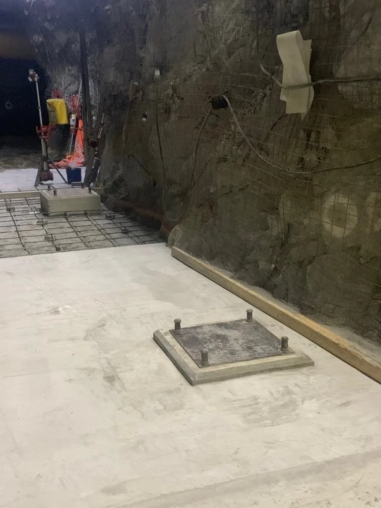 Construction site with a concrete floor, a metal plate with bolts, and a partially built wall with wiring and protective coverings.