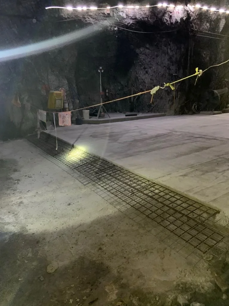 Indoor construction site with concrete floor, metal rebar grid, and safety ropes, surrounded by rocky walls.